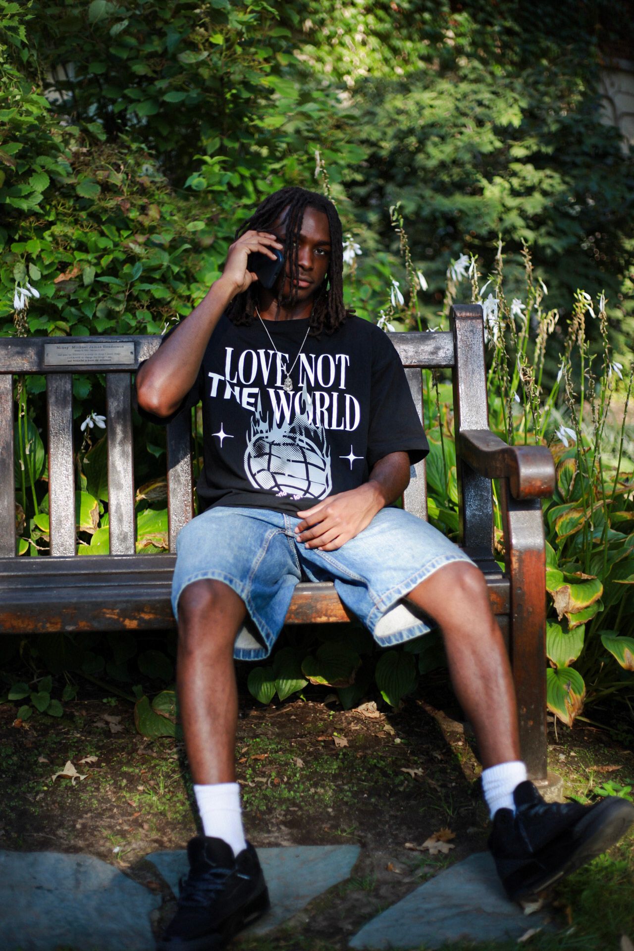 Man in black 'Love Not The World' graphic tee sitting on wooden bench talking on phone outdoors.