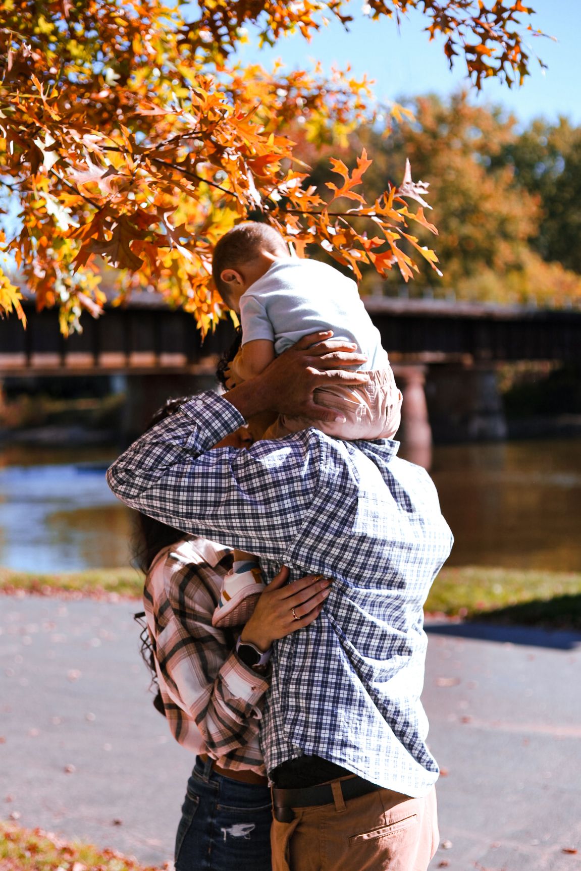 A parent lovingly holds their baby close under vibrant orange autumn leaves near a river bridge.