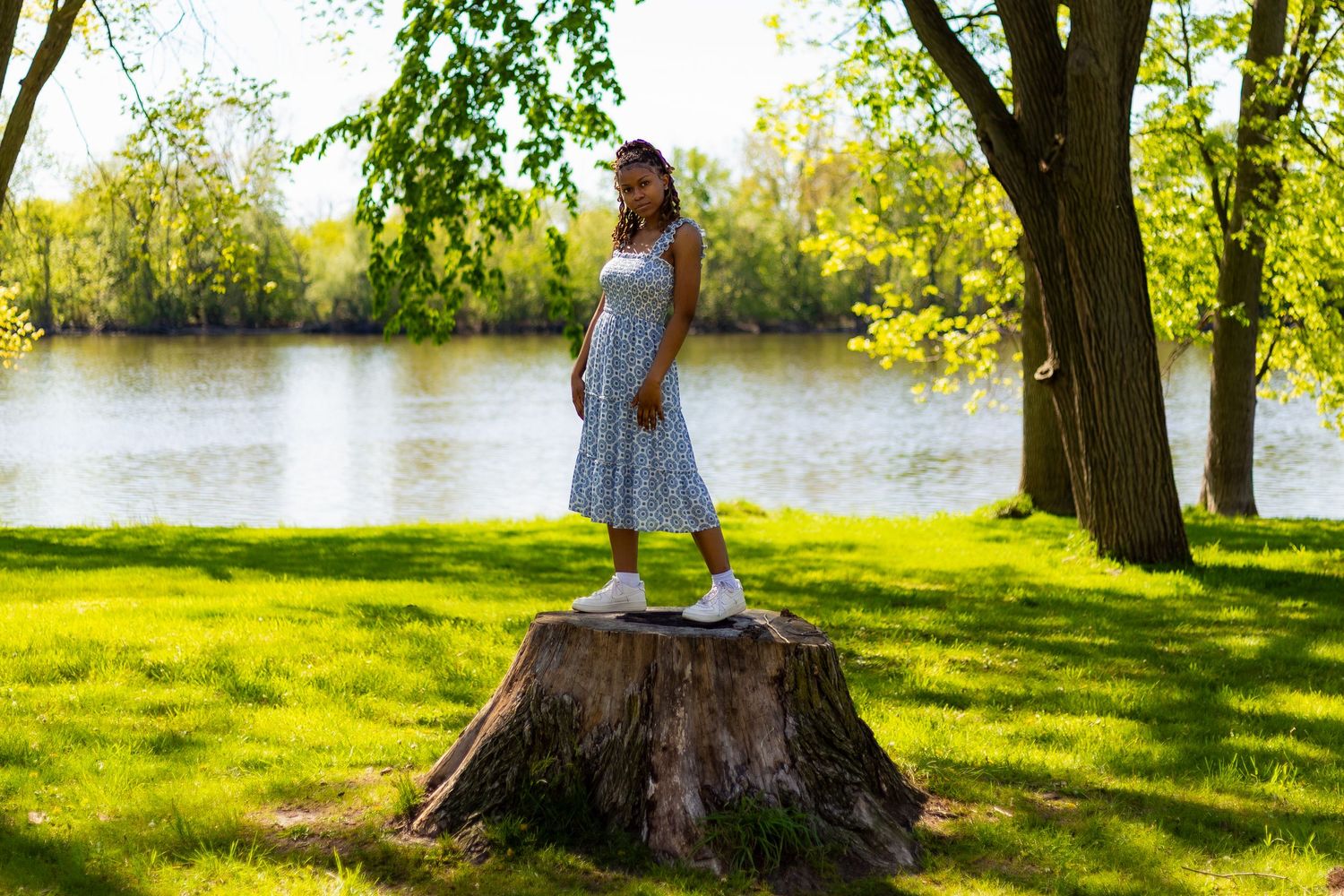 Young Black woman in blue sundress stands on tree stump near lake in spring park.