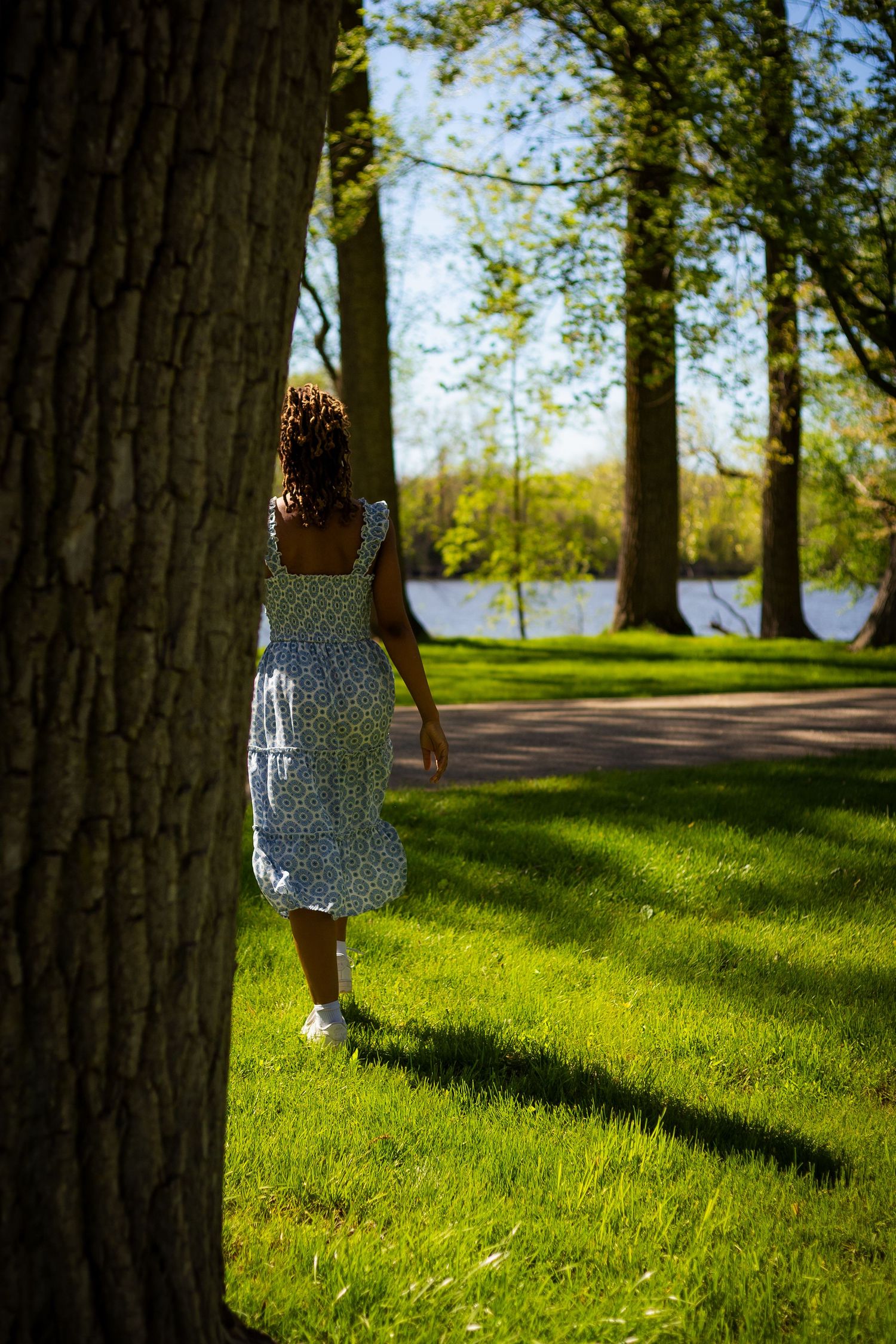 Woman in blue floral sundress walks past large tree trunk in sunny park toward tranquil lake.