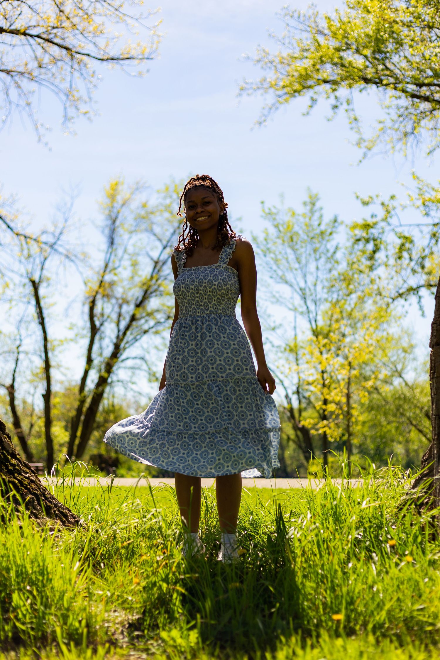 Young Black woman in blue floral dress twirls joyfully on a grassy park path under a bright blue sky.