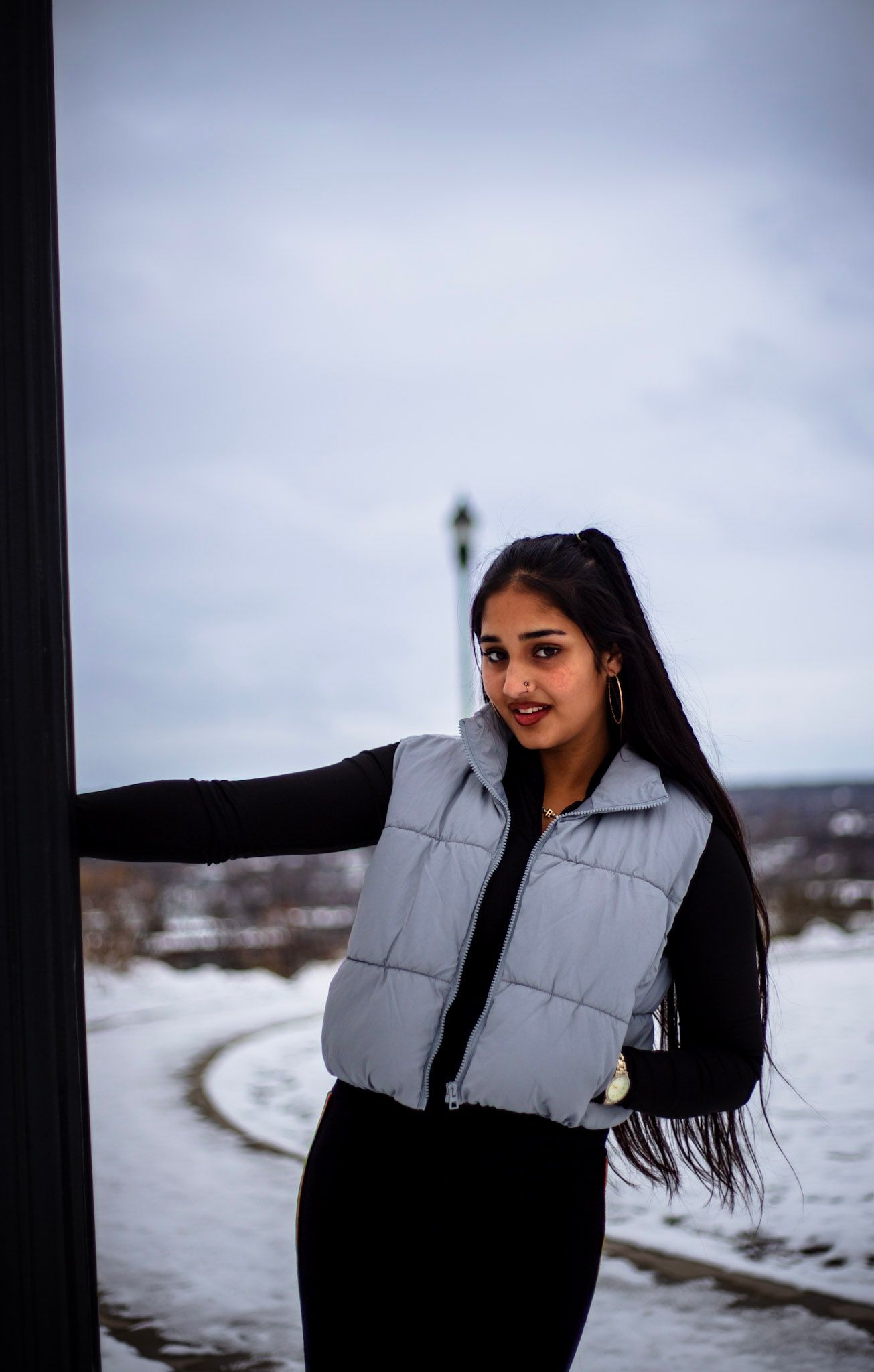 Young woman in silver quilted vest and black outfit poses by dark pillar in snowy winter park setting.