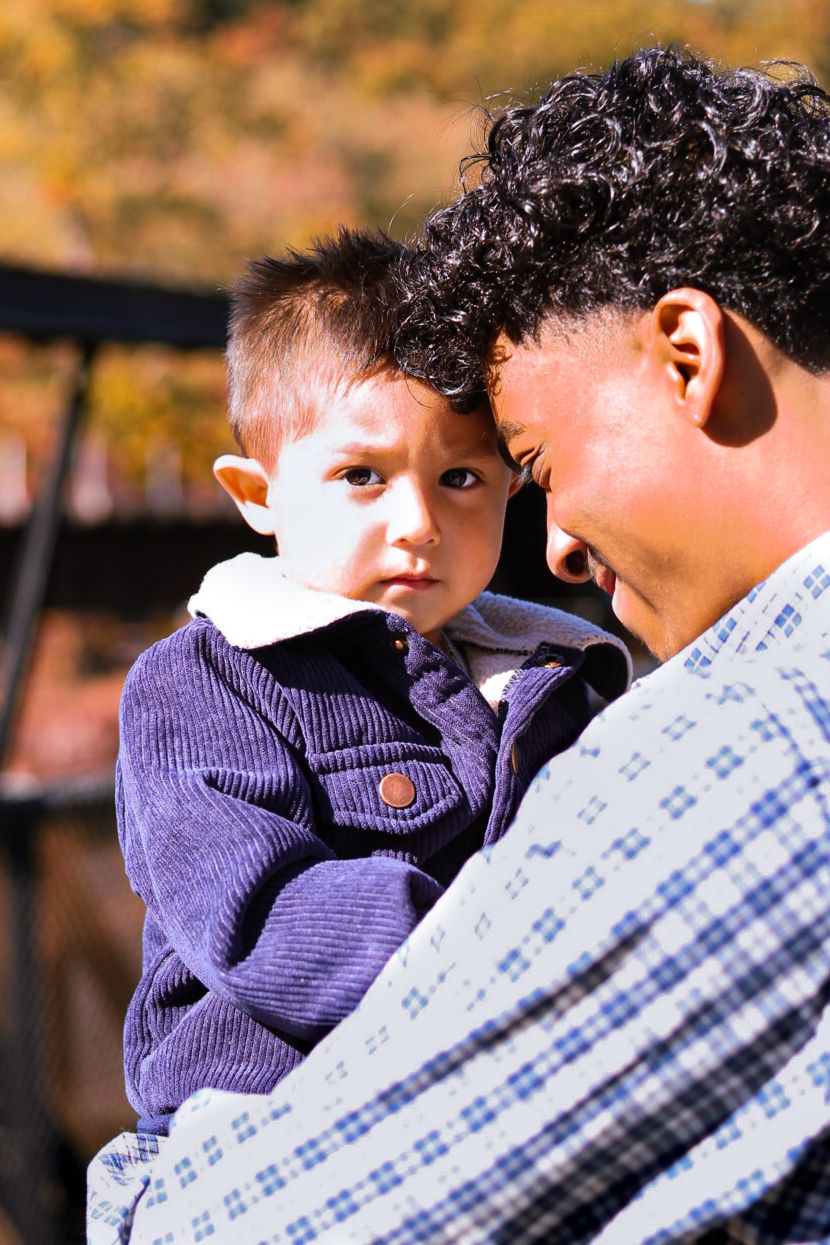 Father holding young toddler son close during fall family portrait session with autumn foliage.