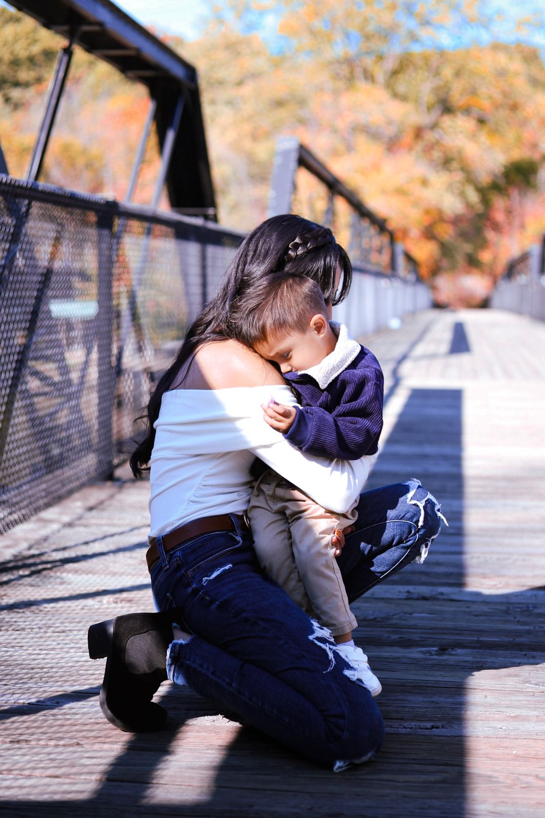 Mother kneeling on bridge hugging young toddler son during autumn family photo session outdoors.