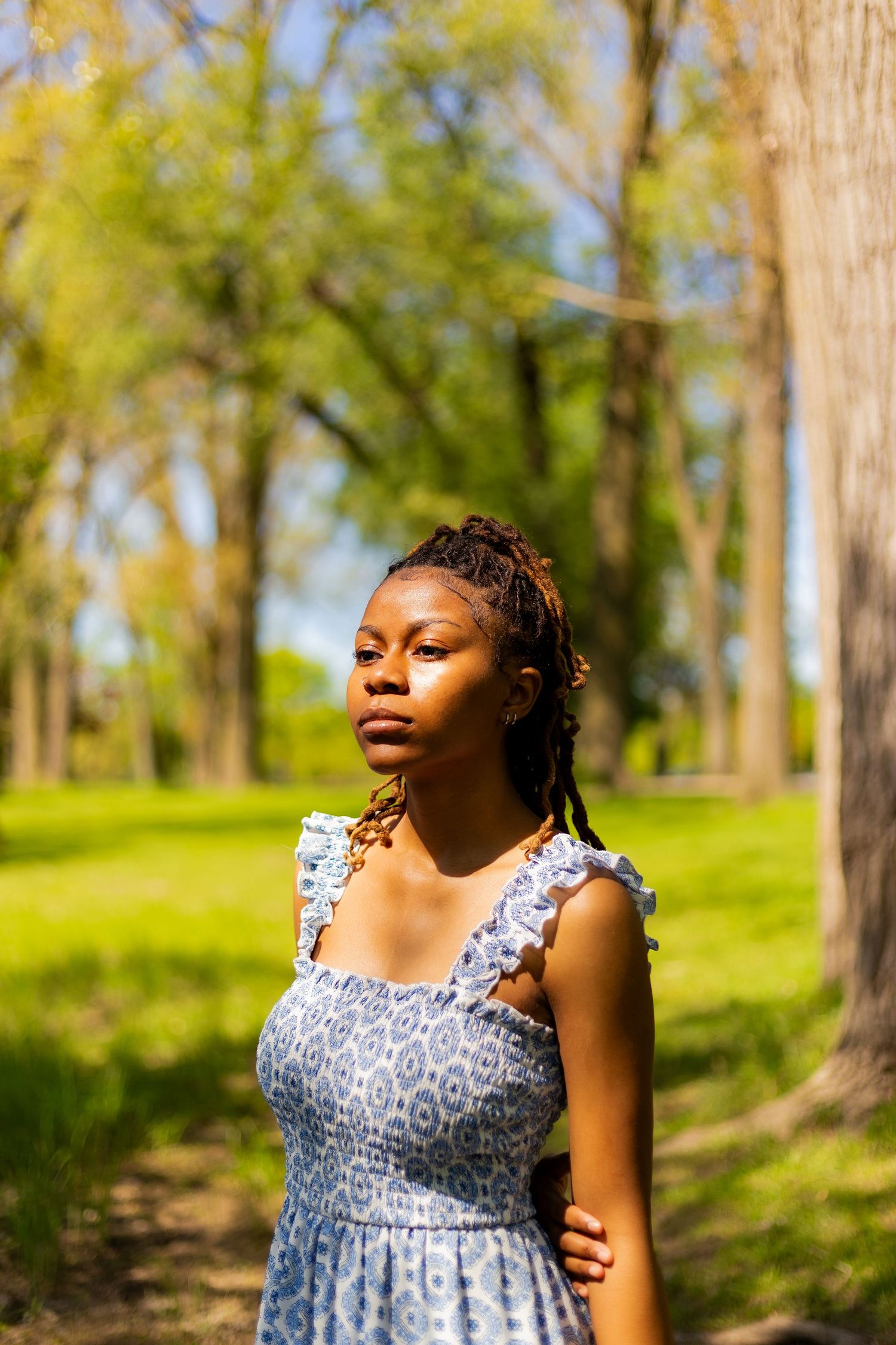 Young Black woman with locs in a blue sleeveless dress stands confidently in a lush green park.