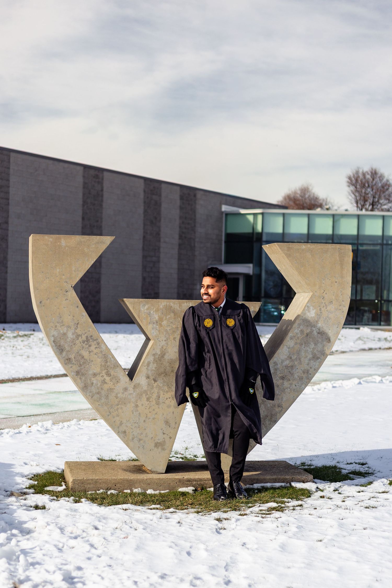 Graduate in black cap and gown poses before concrete university sculpture in snowy winter campus setting.