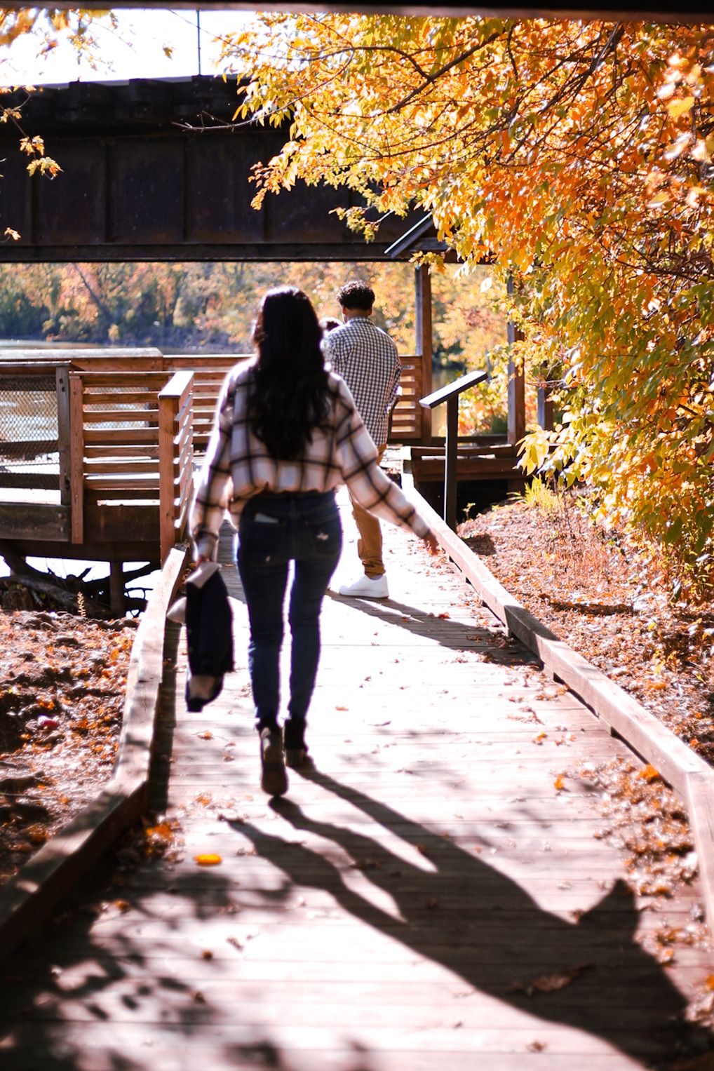 Woman walking along a wooden boardwalk path surrounded by golden autumn leaves toward a bridge.