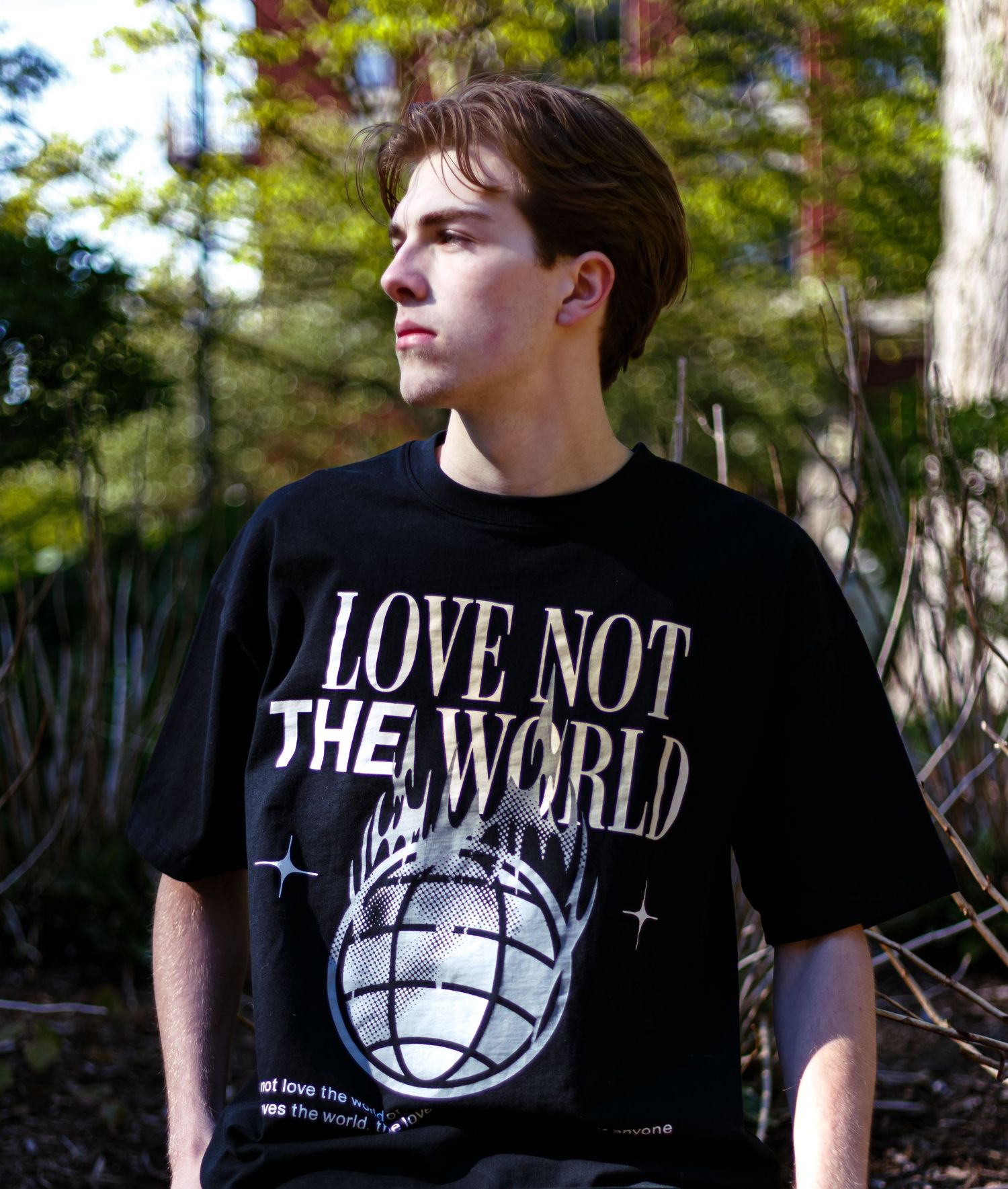 Young man wearing a black 'Love Not The World' graphic t-shirt standing outdoors among trees.