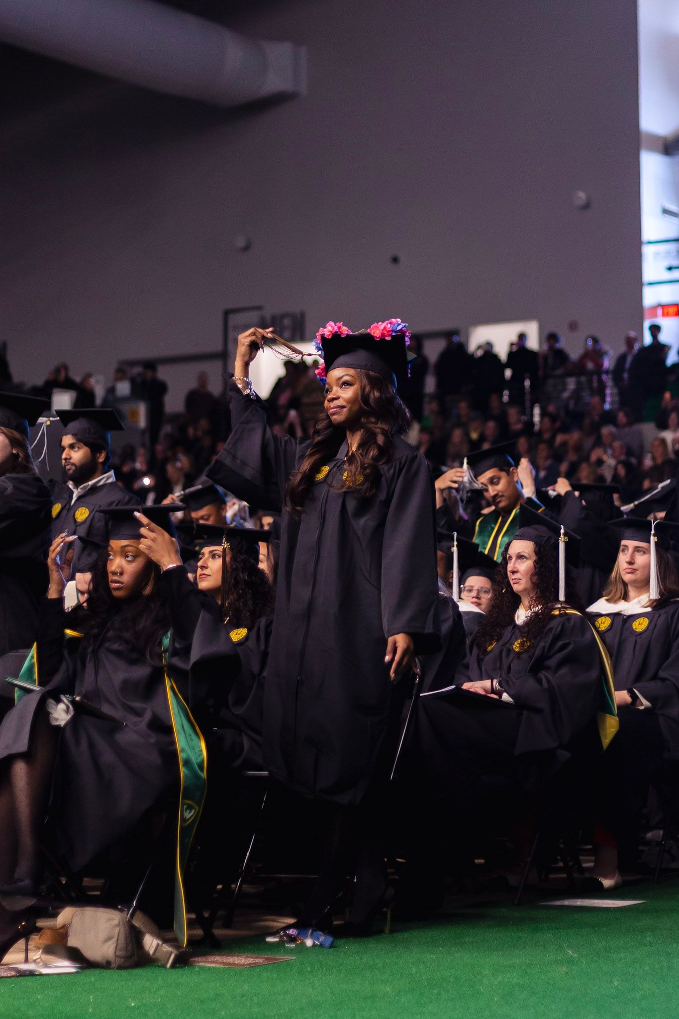 A graduate in a floral crown adjusts her cap during a university commencement ceremony indoors.
