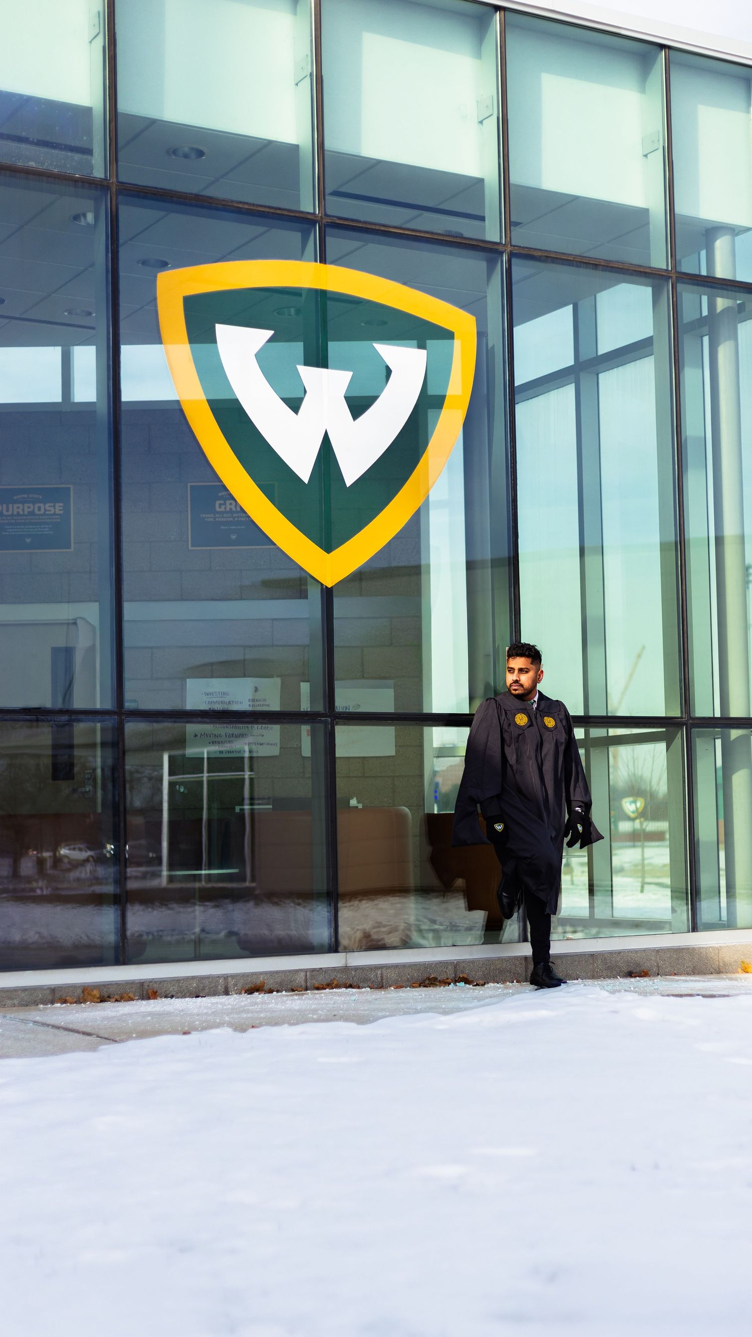 Graduate in commencement regalia walking past Wayne State University W shield logo on glass facade.
