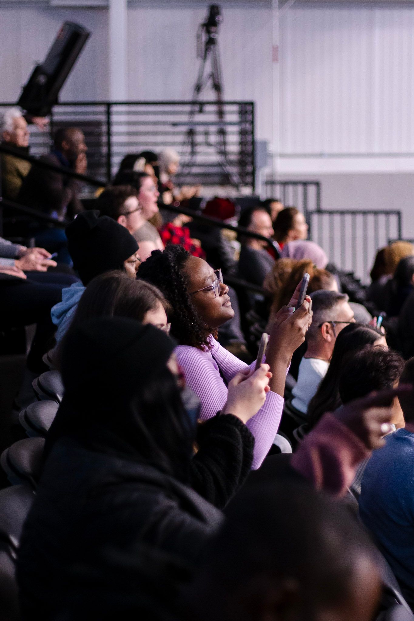 Diverse audience members seated in bleachers at an indoor event, with a young woman in purple holding a phone.