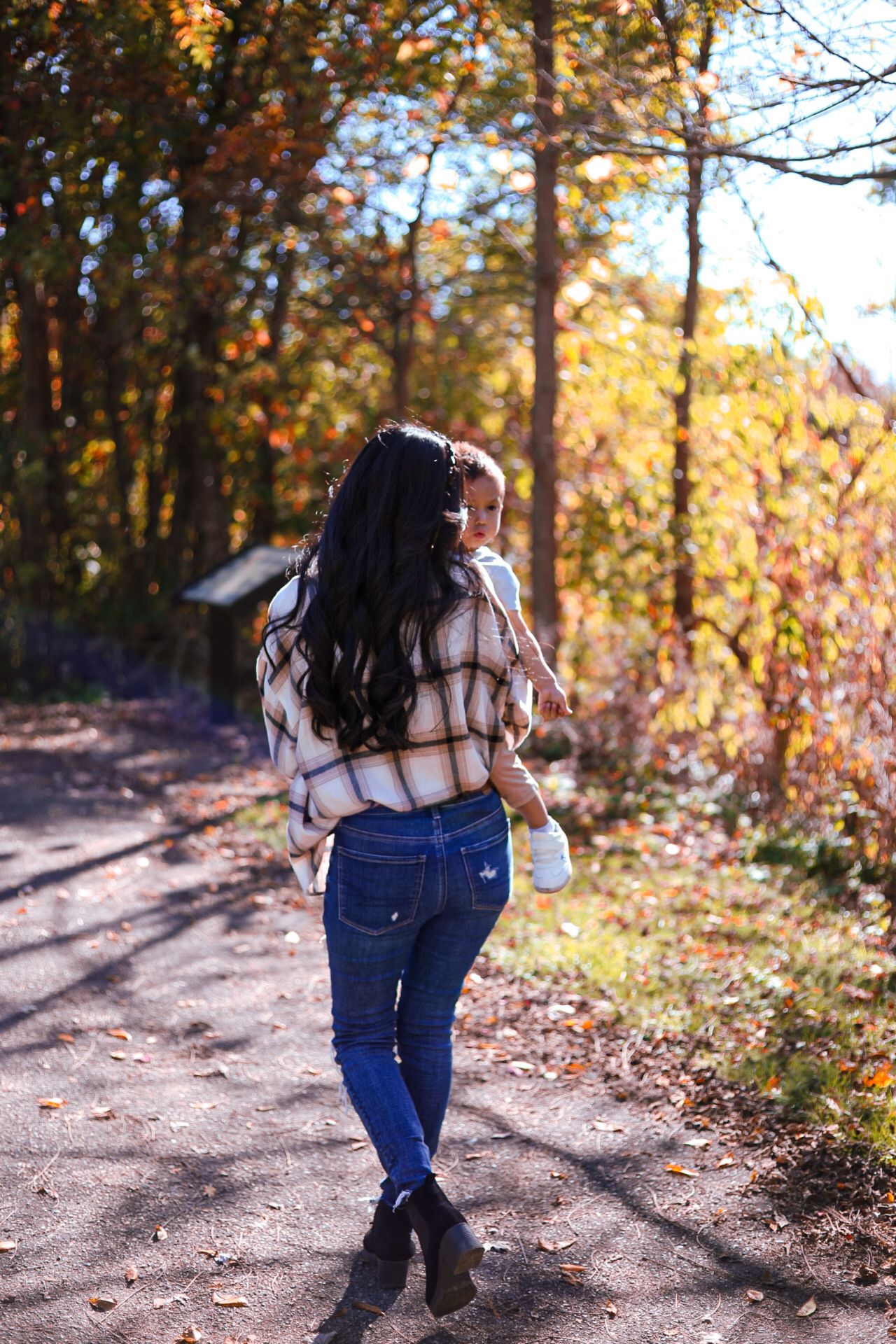 Woman in plaid shirt carrying toddler while walking along a sunlit autumn forest path.