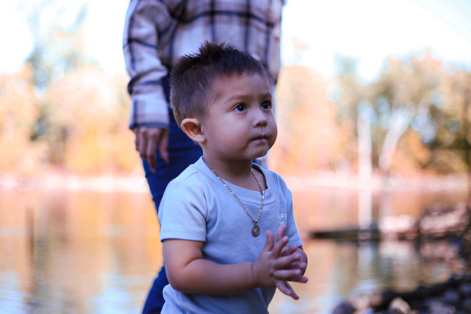 A young toddler with a gold necklace stands near a river with autumn trees in the background.