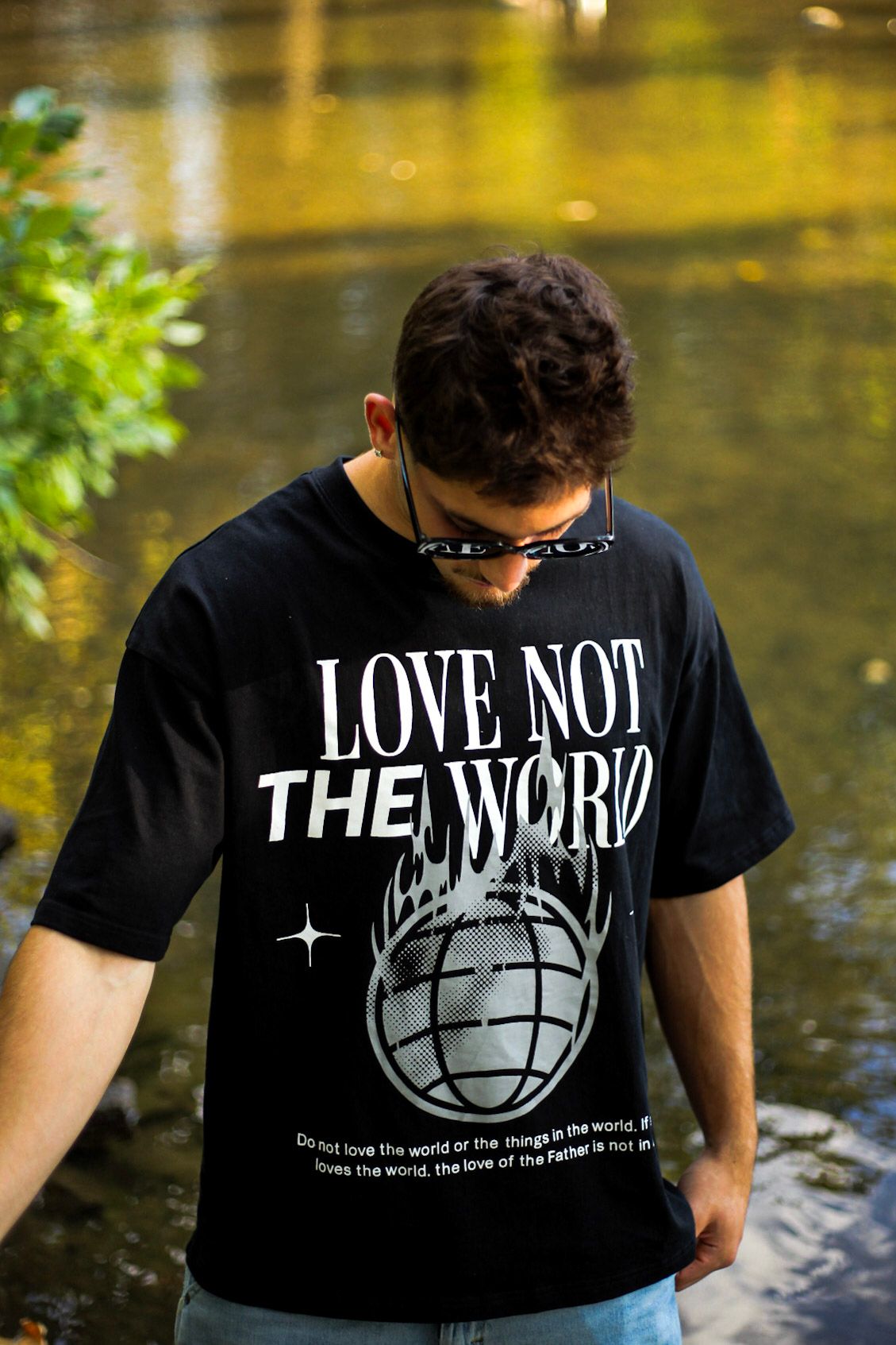 Man looking down wearing black Love Not The World graphic t-shirt near a scenic river.