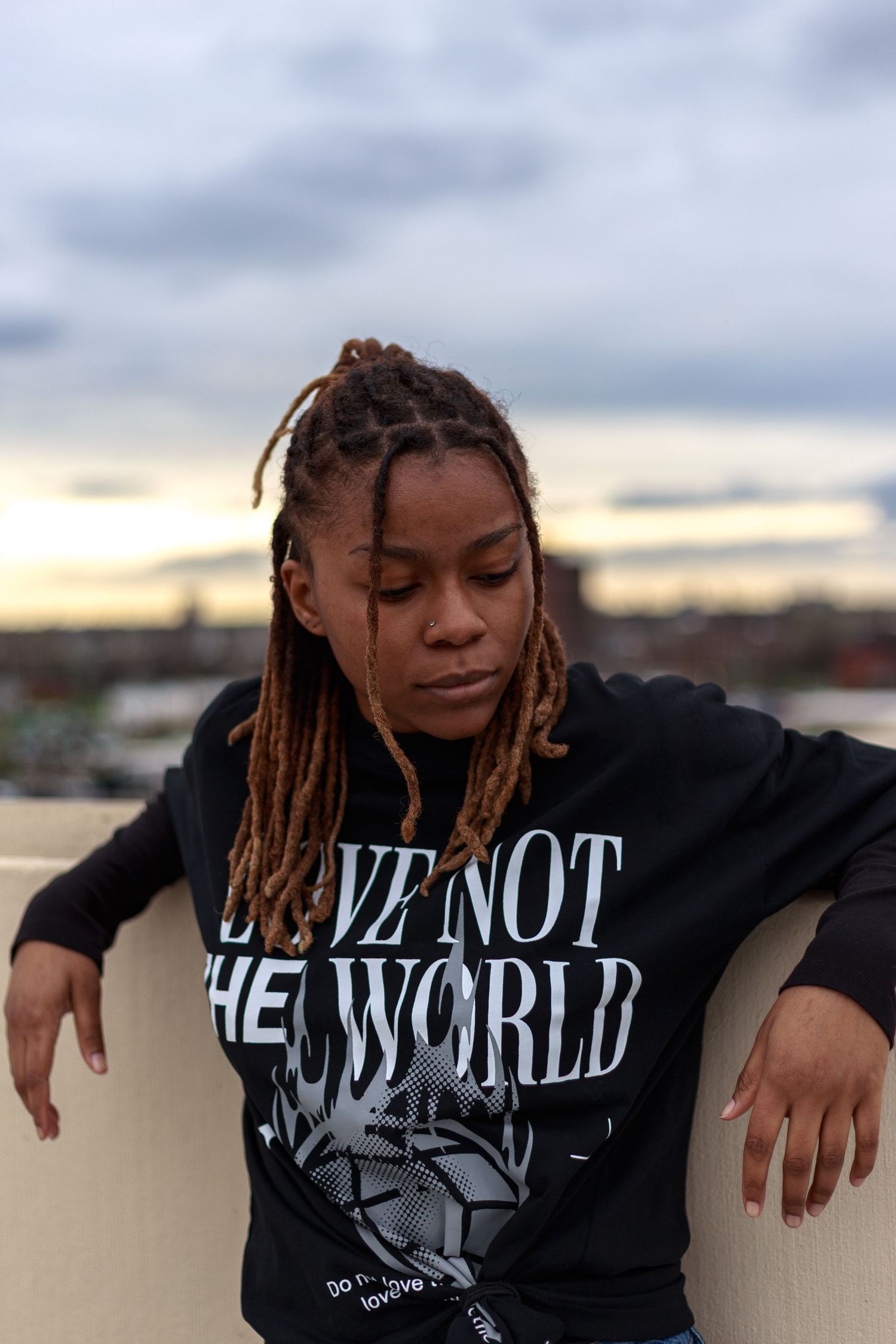 Young woman with locs in a black Love Not The World sweatshirt poses pensively on a rooftop at dusk.