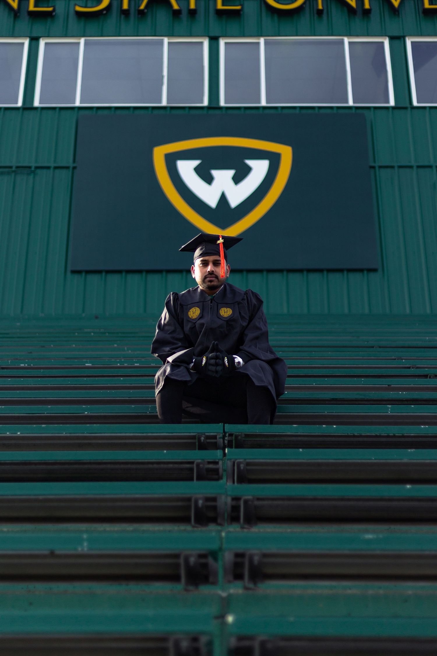 Wayne State University graduate sits in green bleachers beneath the iconic WSU logo.