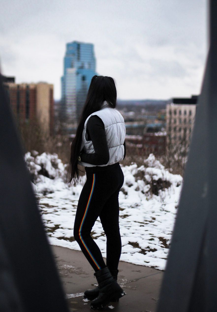 Young woman in white puffer vest stands overlooking snowy urban cityscape with tall buildings.