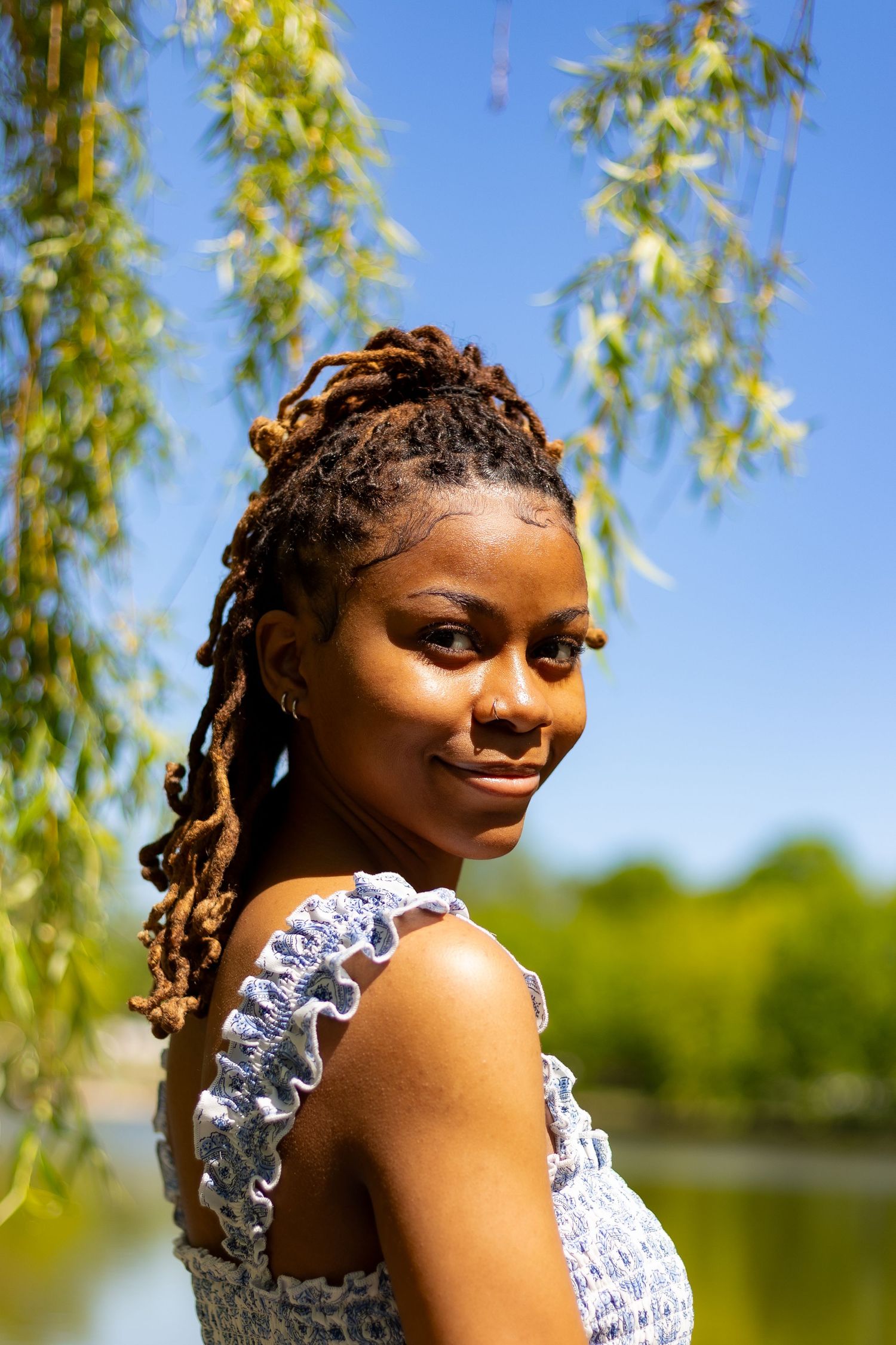 Smiling young Black woman with dreadlocks in blue sundress stands by lake under willow tree.