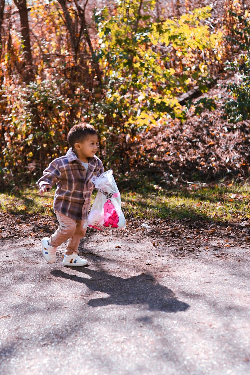 Young toddler in plaid jacket carrying plastic bag on sunny autumn day outdoors.