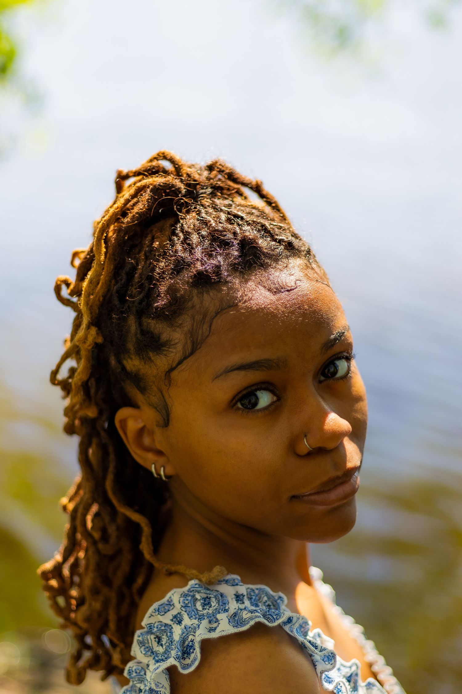 Close-up portrait of a young Black woman with styled locs and hoop earring in a blue floral dress outdoors.