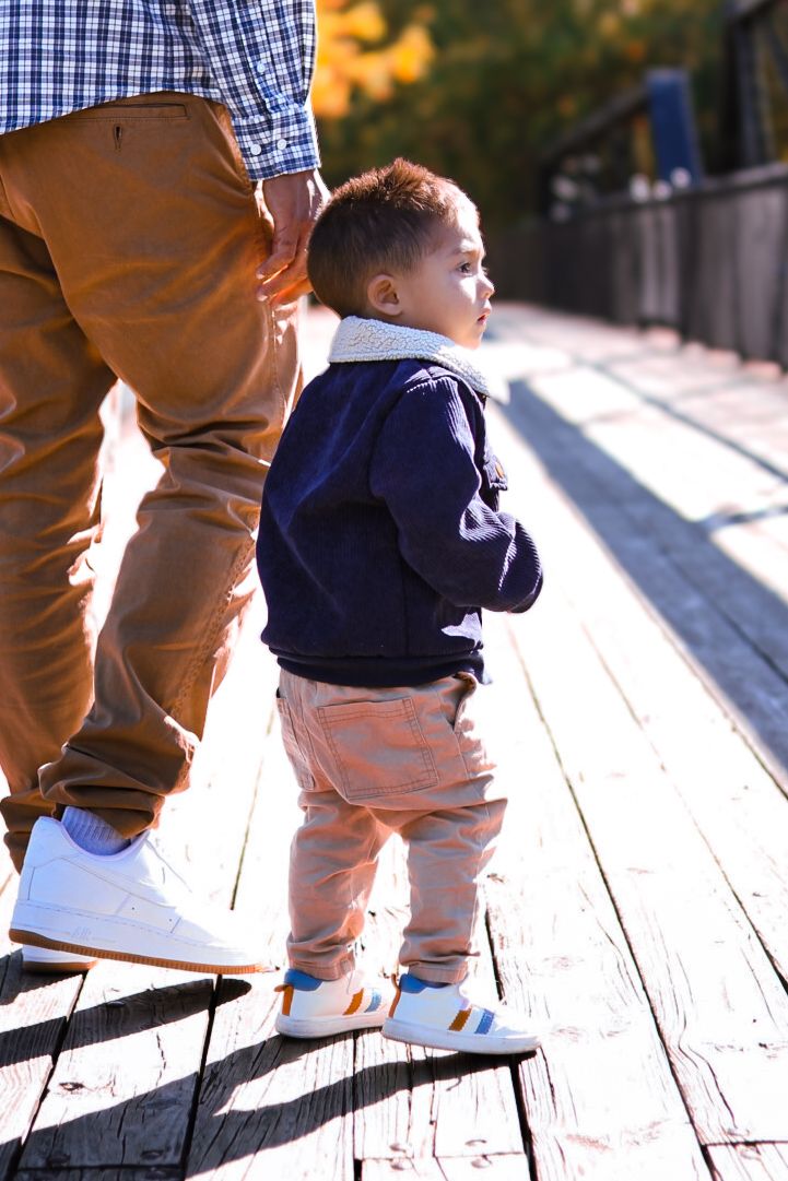 Close-up of toddler in navy jacket and tan pants standing on wooden bridge holding parent's hand in autumn.