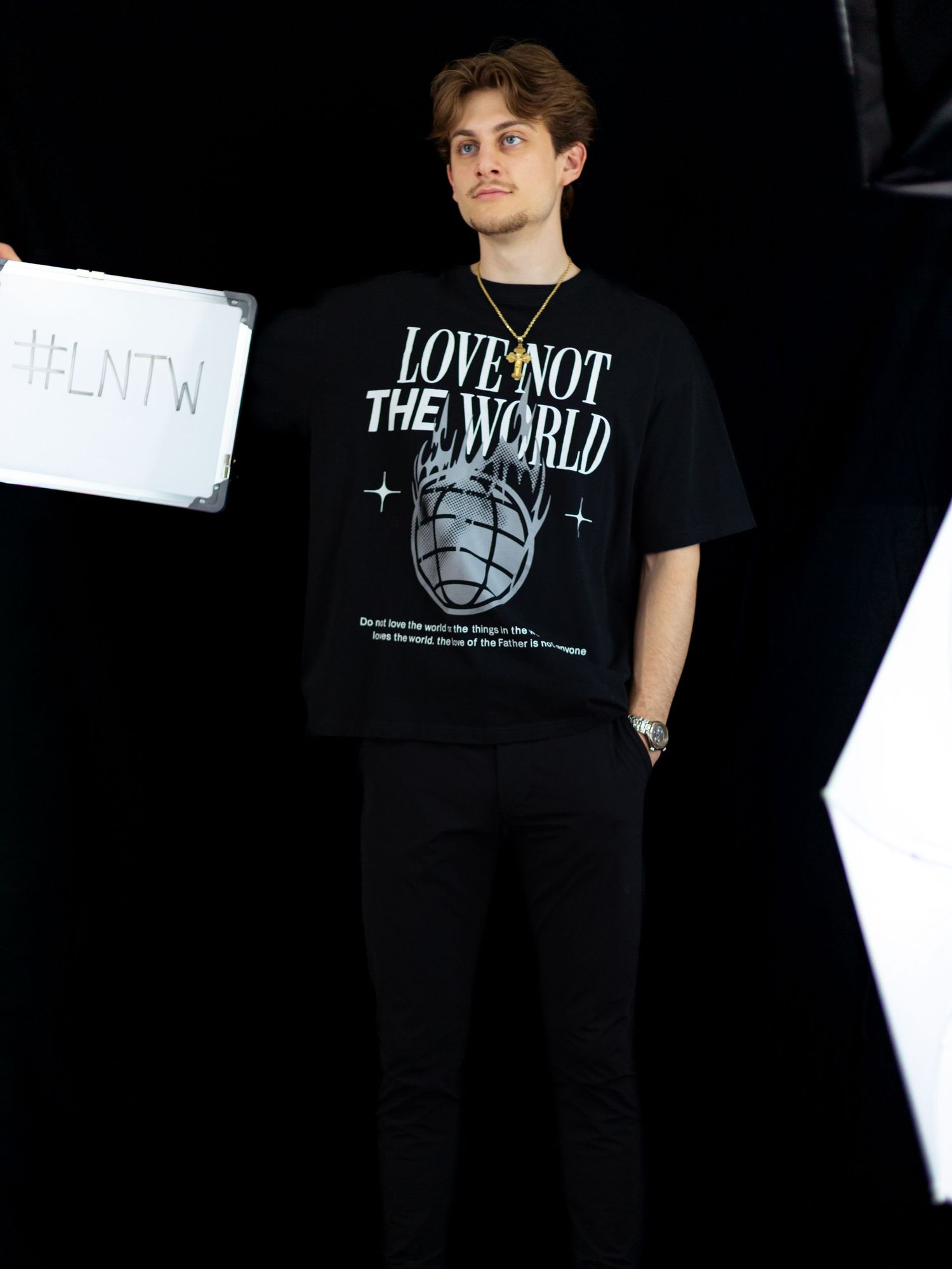 Full-length shot of man holding #LNTW whiteboard in black Love Not The World t-shirt and black jeans.