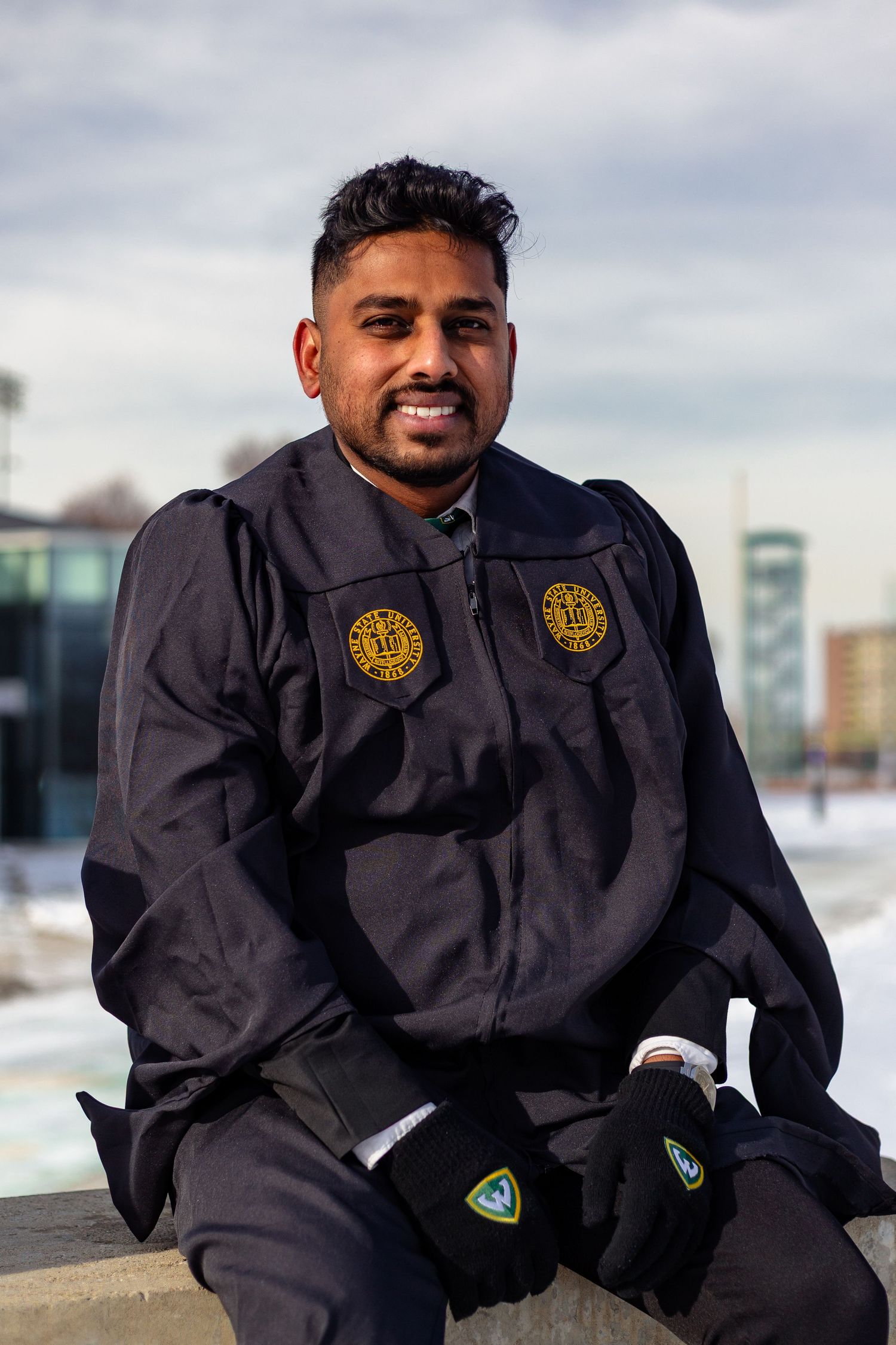 Smiling Wayne State University graduate in navy cap and gown seated outdoors on winter campus.