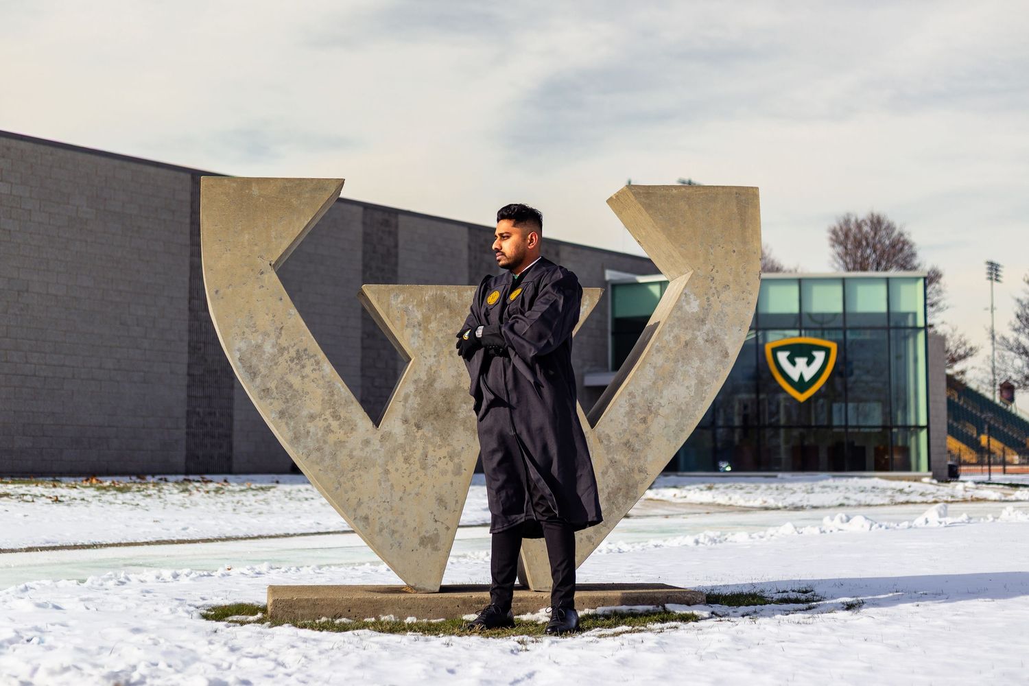 Graduate in black gown posing by Wayne State University sculpture in winter snow.