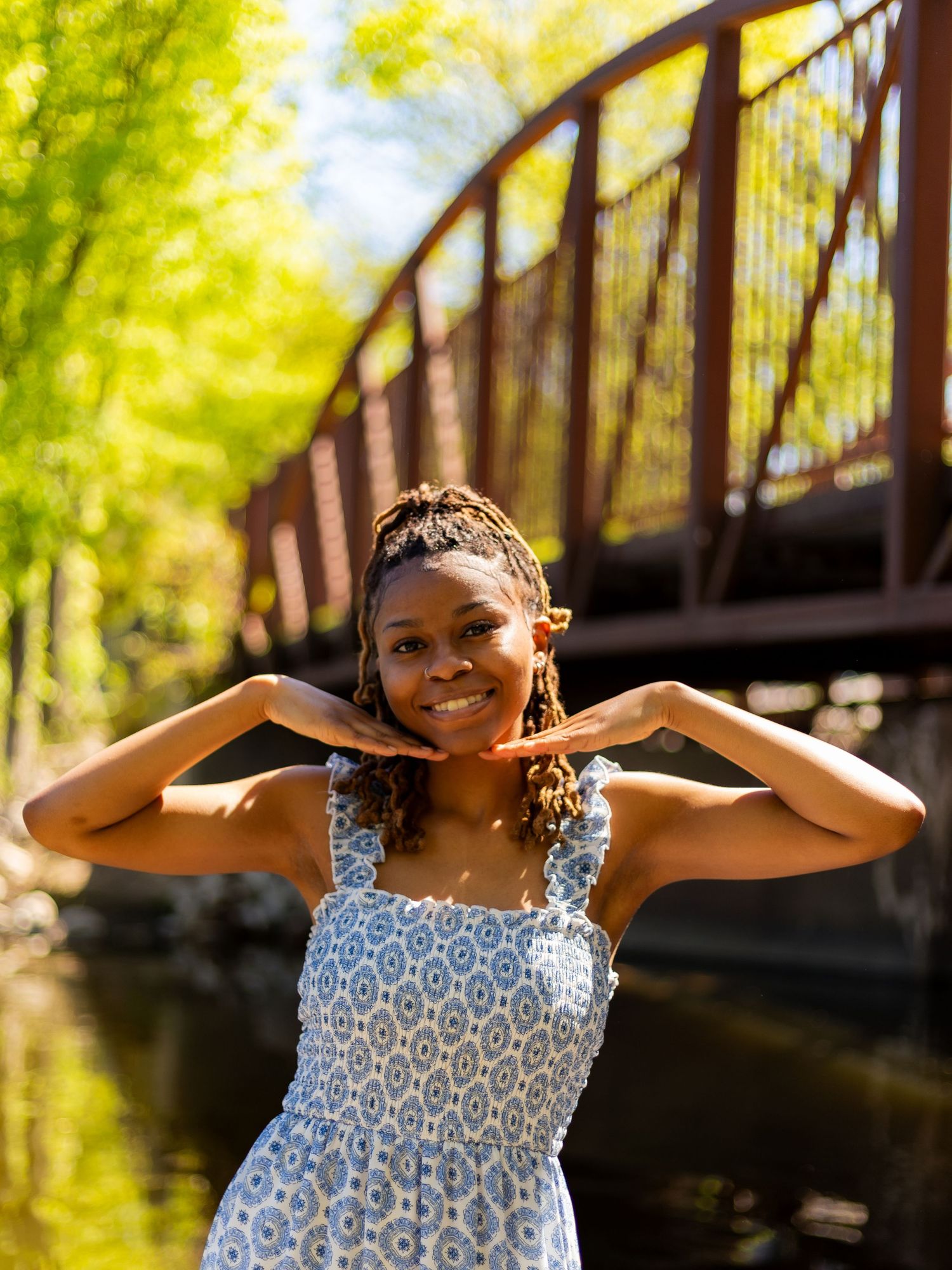 Young Black woman in blue floral sundress smiling near rustic bridge and river on sunny day.