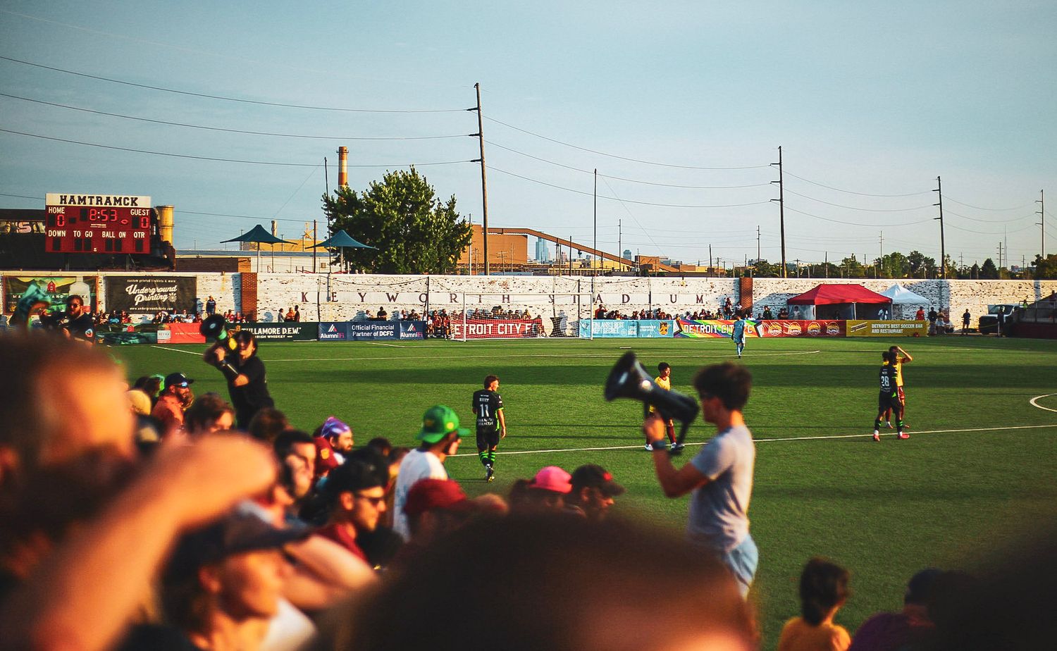 Detroit City FC fans pack the sidelines cheering during a sunny outdoor soccer match at Keyworth Stadium.