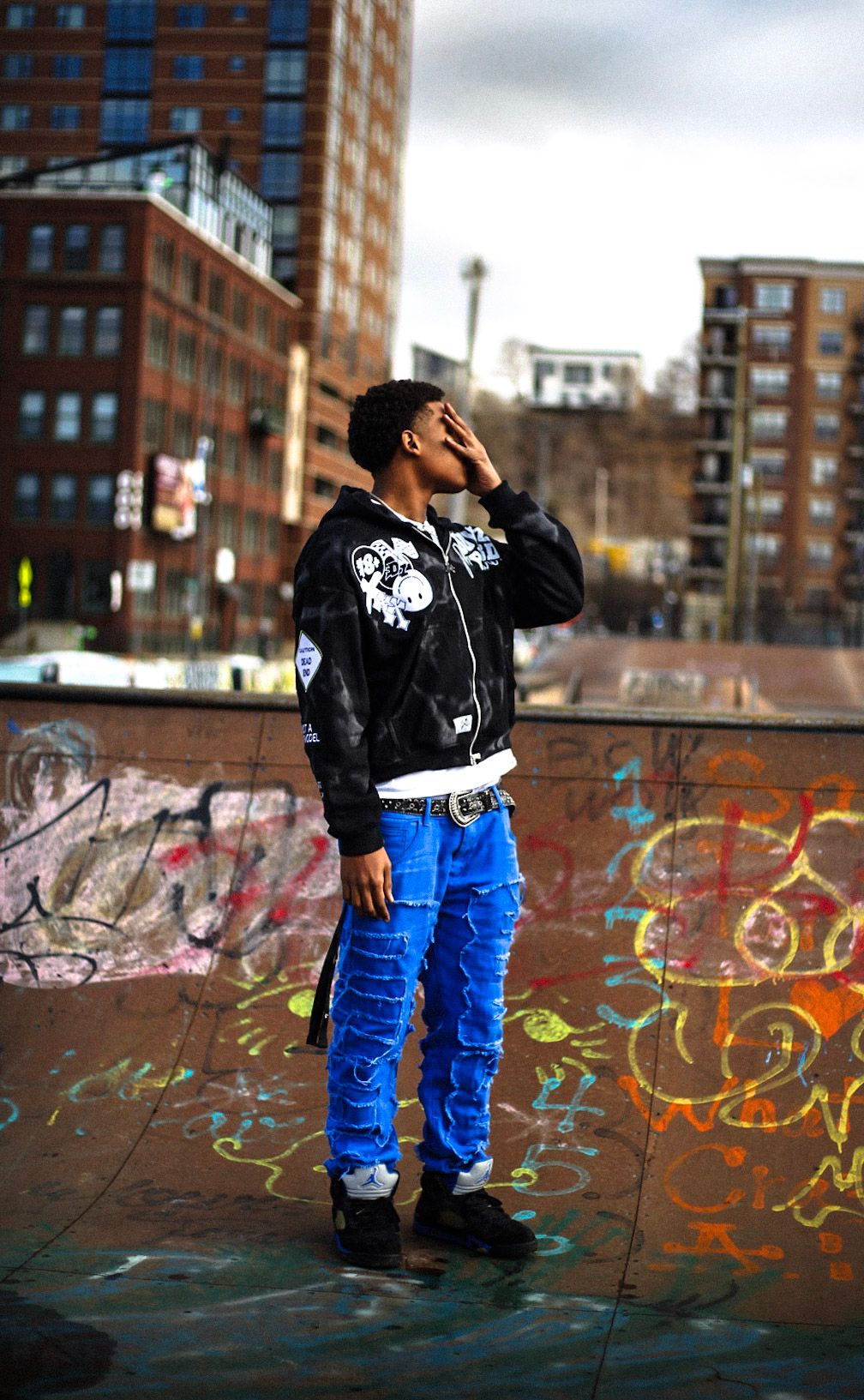 Young man in blue pants and black jacket shielding eyes while posing at graffiti skate park.