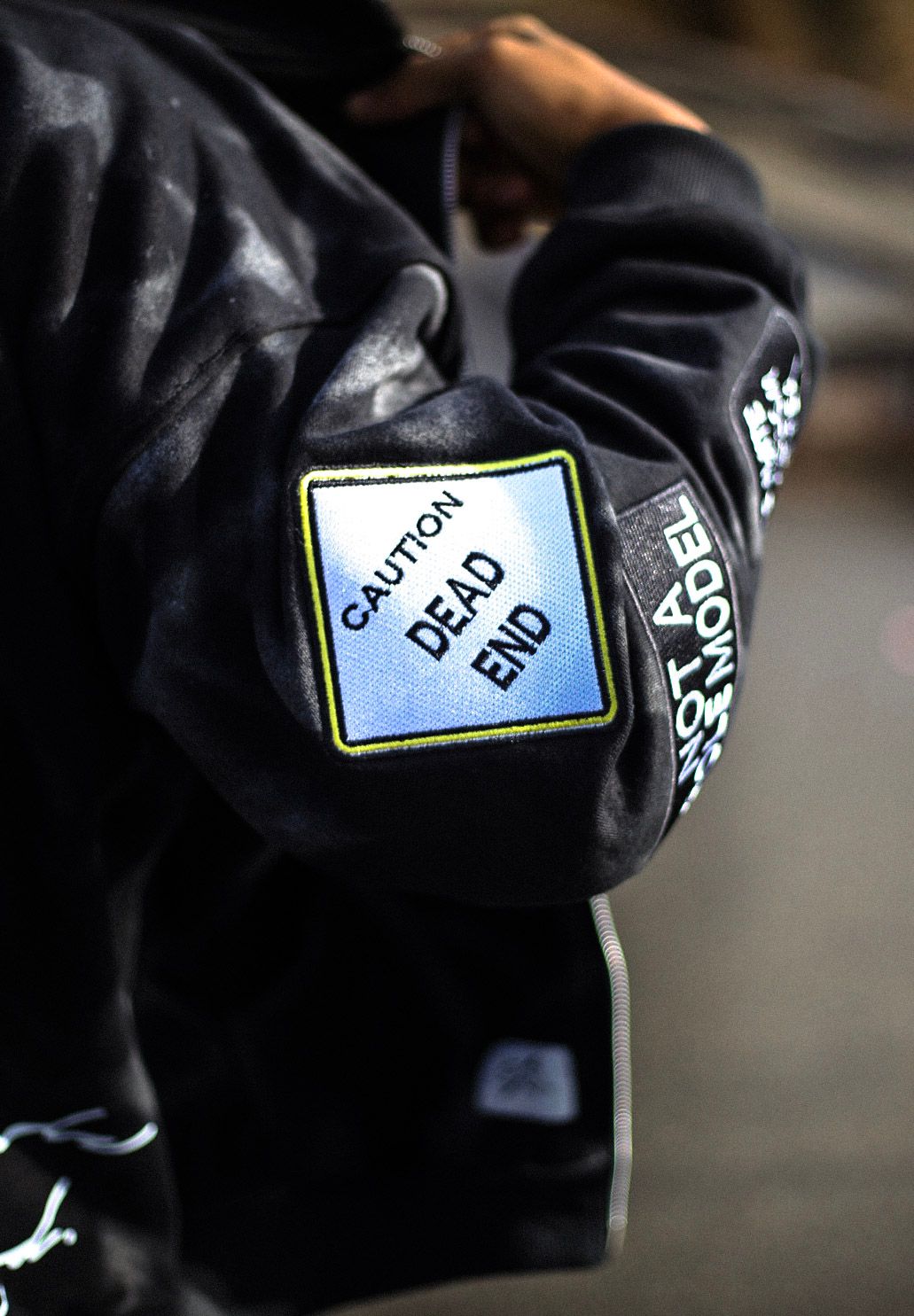 Black jacket sleeve featuring embroidered Caution Dead End street sign patch detail.