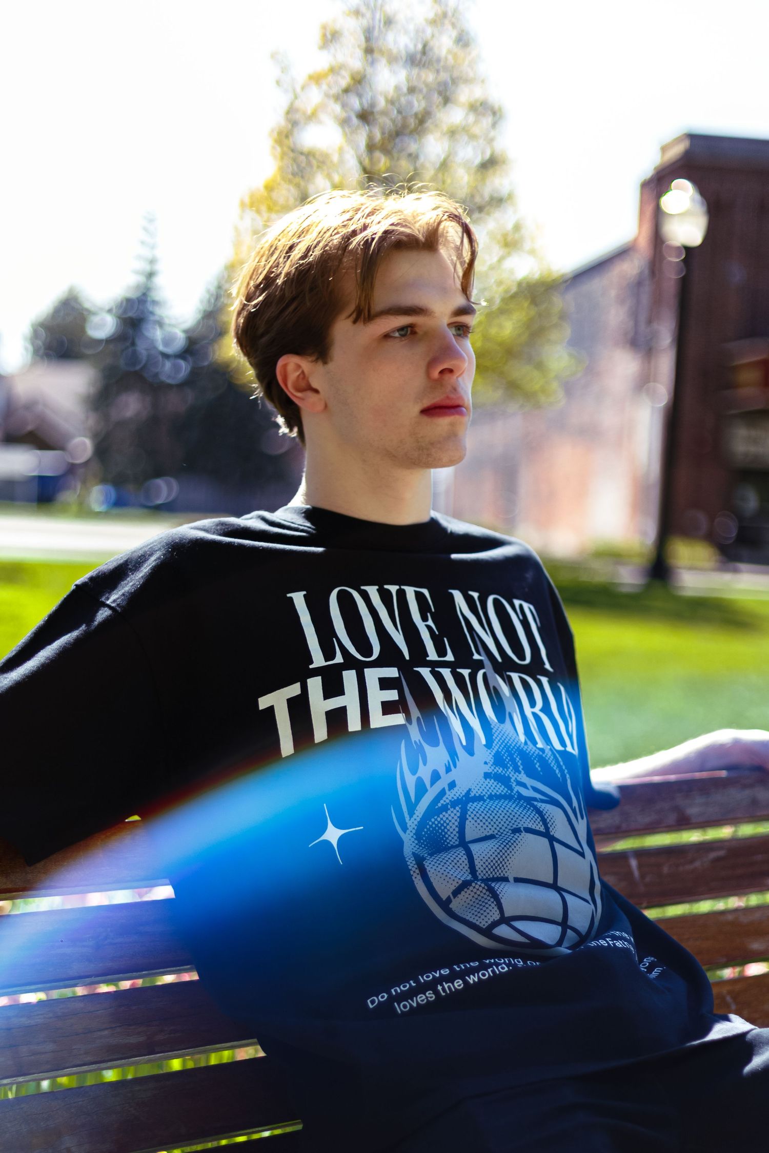 Close-up of young man on bench wearing black Love Not The World oversized graphic tee in sunlight.