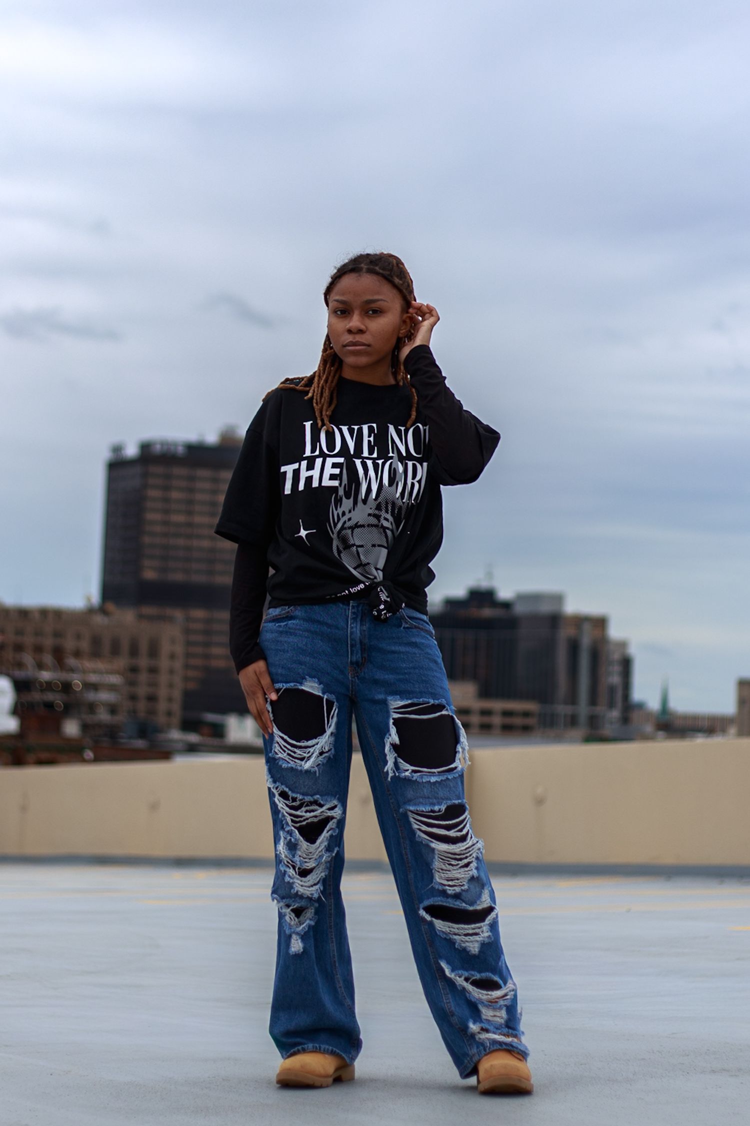 Woman with locs stands on rooftop in ripped wide-leg jeans and black 'Love Not The World' tee with city skyline.
