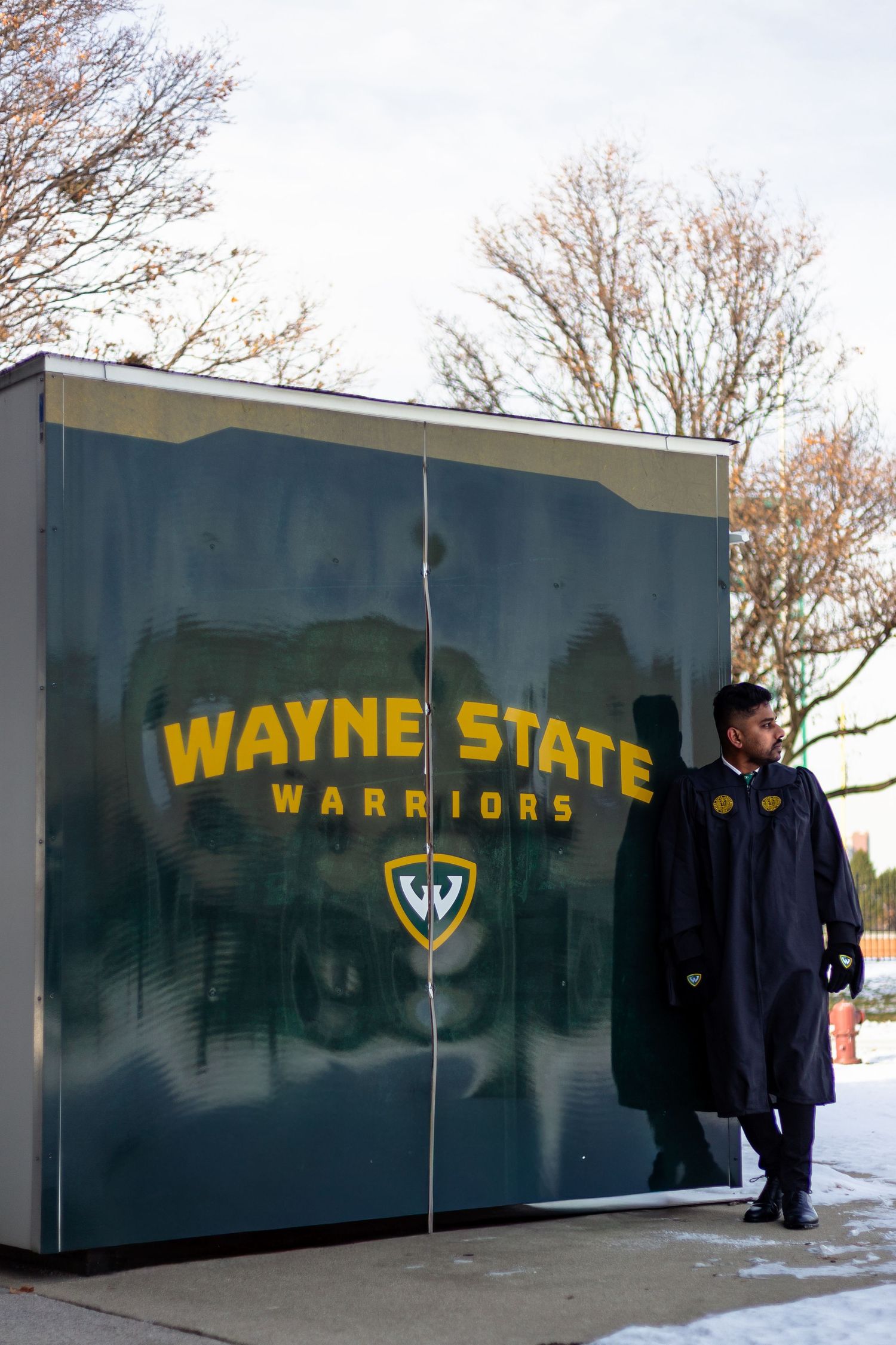 Wayne State Warriors graduate posing beside green branded display wall in graduation regalia.