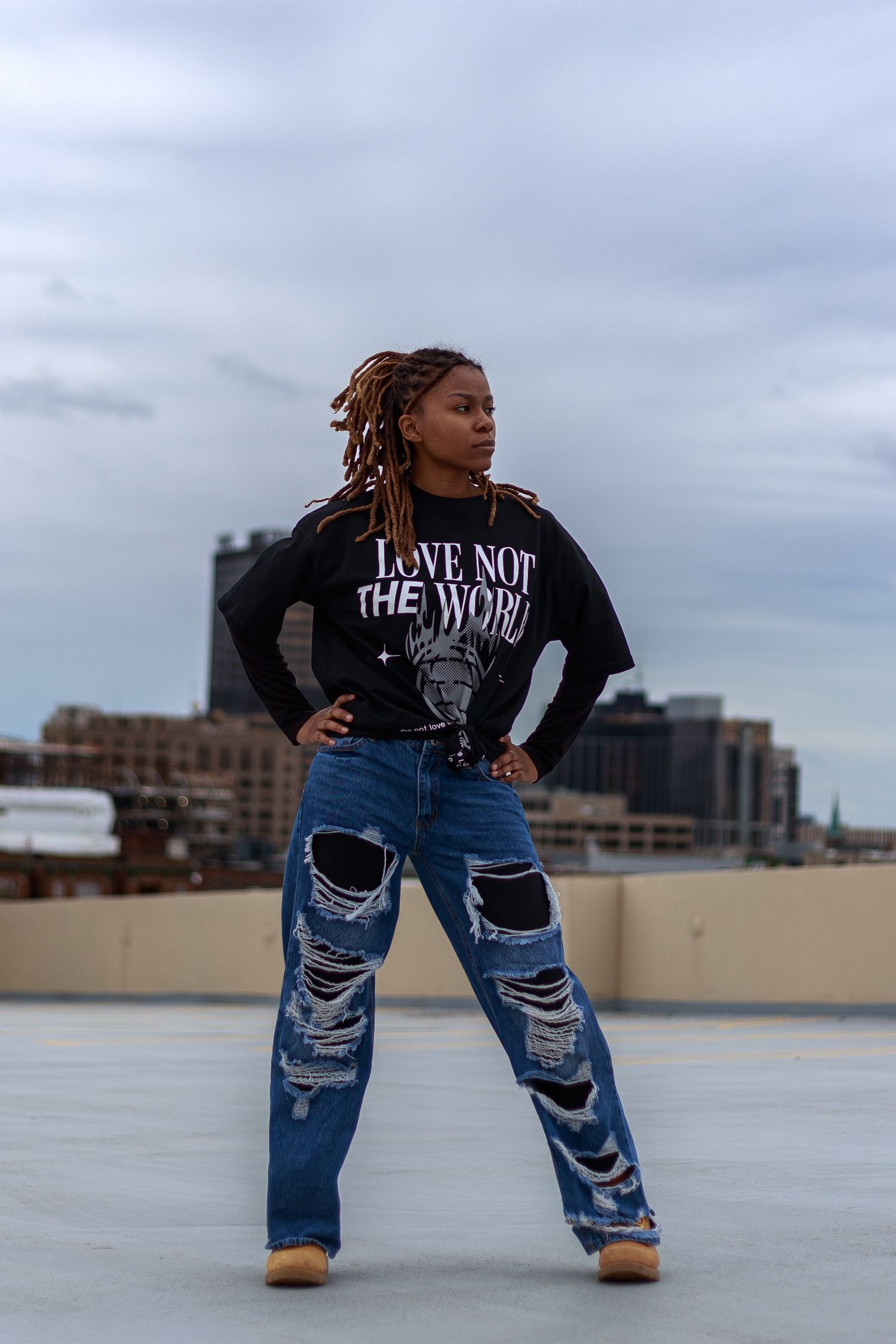 Confident woman in distressed wide-leg jeans and black graphic tee poses with hands on hips on a rooftop.