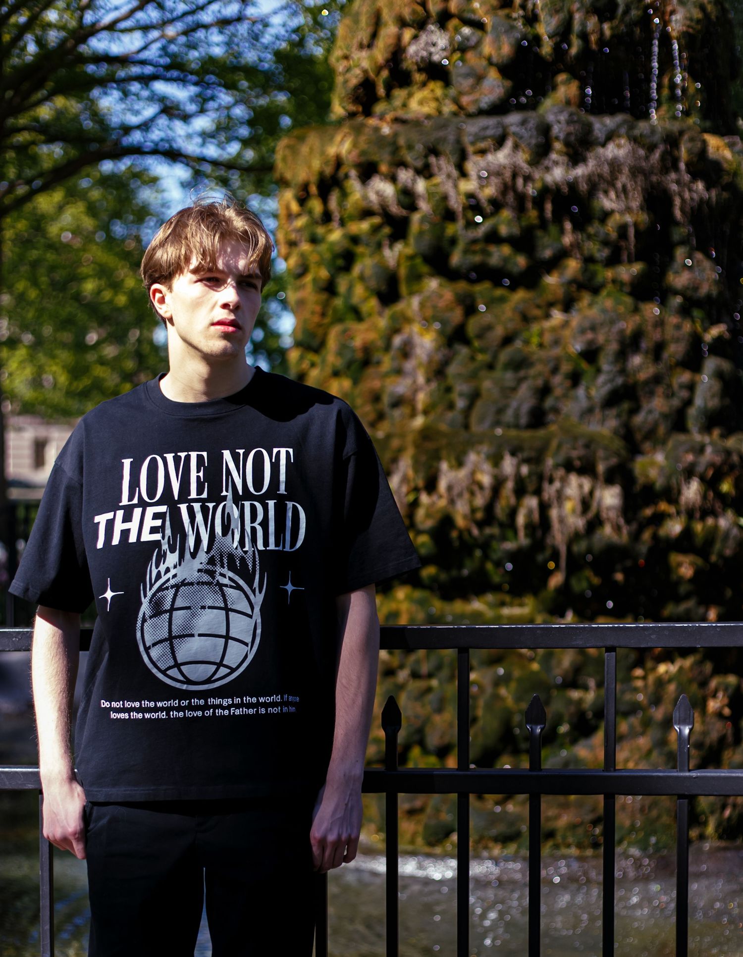 Young man standing near fountain wearing black Love Not The World graphic tee in natural setting.