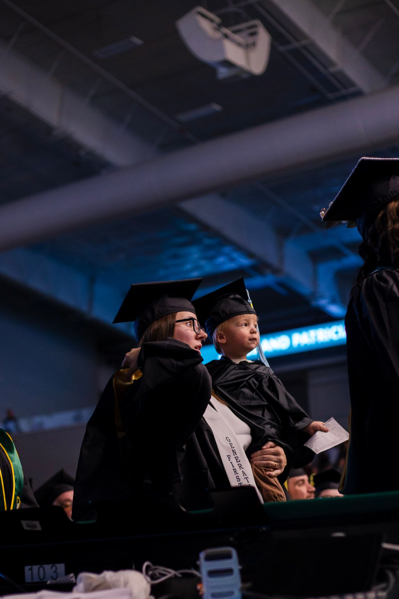 Graduate in black cap and gown holding a young child dressed in a mini graduation gown at a commencement ceremony.