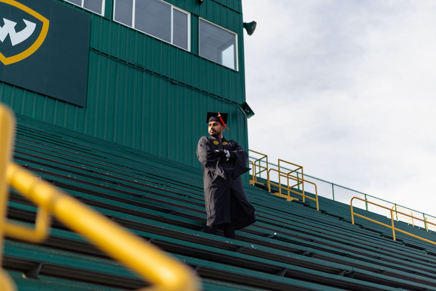 Wayne State University graduate stands in green stadium bleachers wearing black graduation regalia.