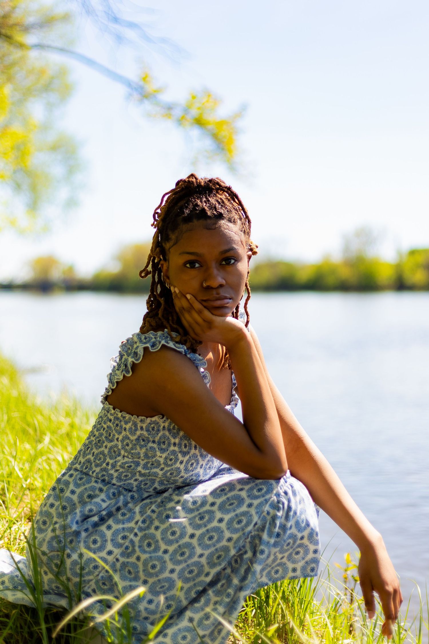 Young Black woman in blue floral dress sits gracefully by calm lake in sunny park.