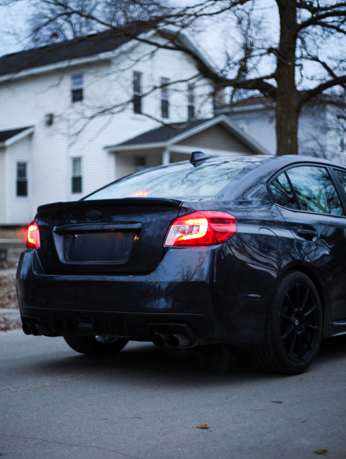 Rear view of a dark Subaru WRX sedan with glowing red taillights parked on a residential street.