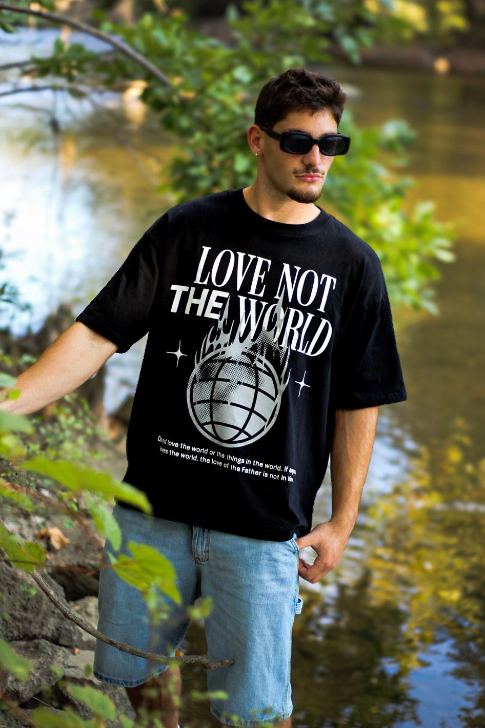 Man in sunglasses wearing Love Not The World black graphic tee standing near a woodland stream.