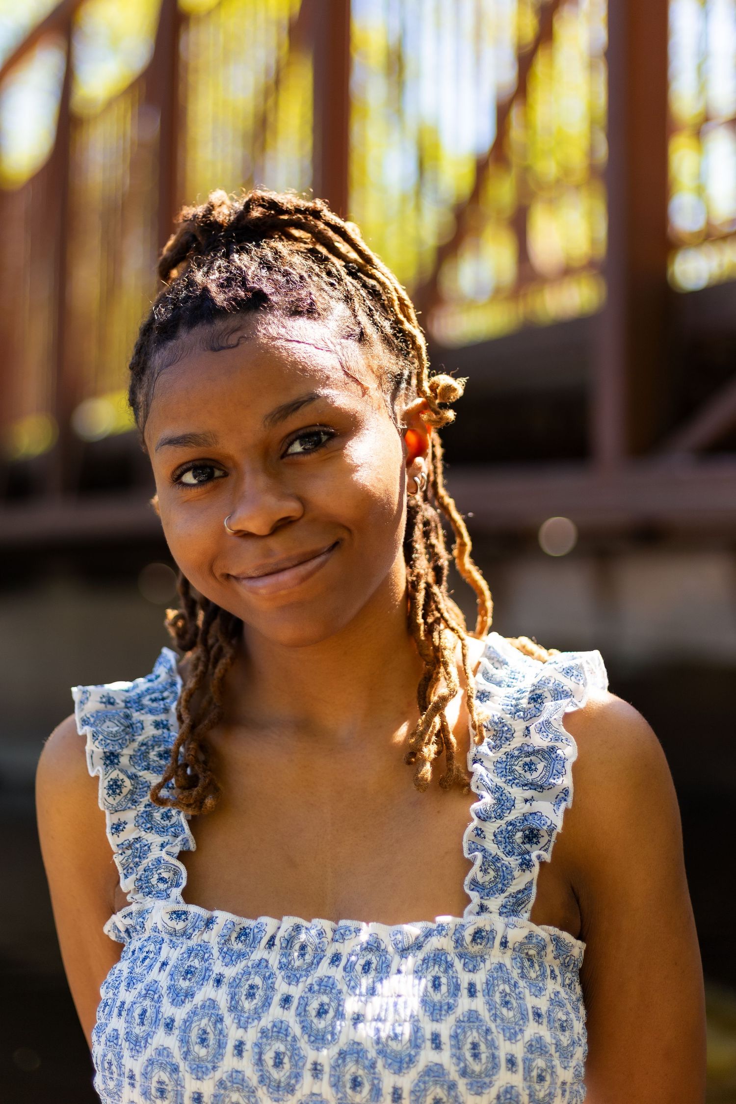 Young Black woman with braided locs smiling softly outdoors near a rustic bridge in a blue floral sundress.