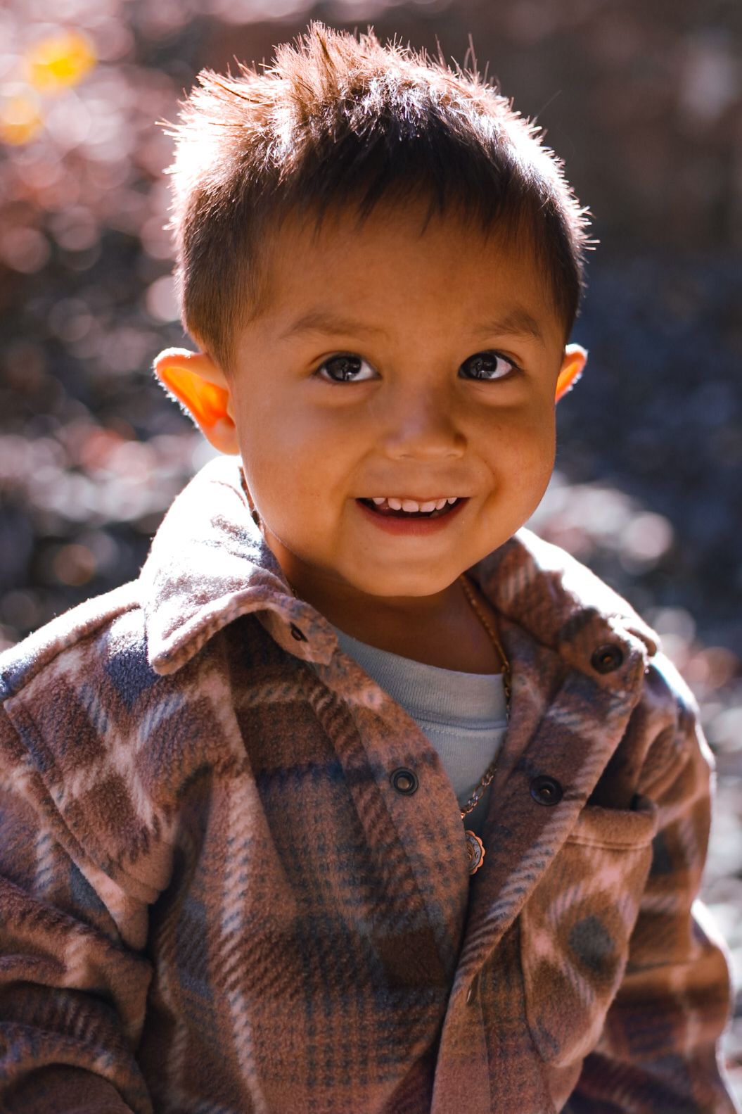 Smiling young boy in plaid flannel shirt poses in warm sunlight outdoors.