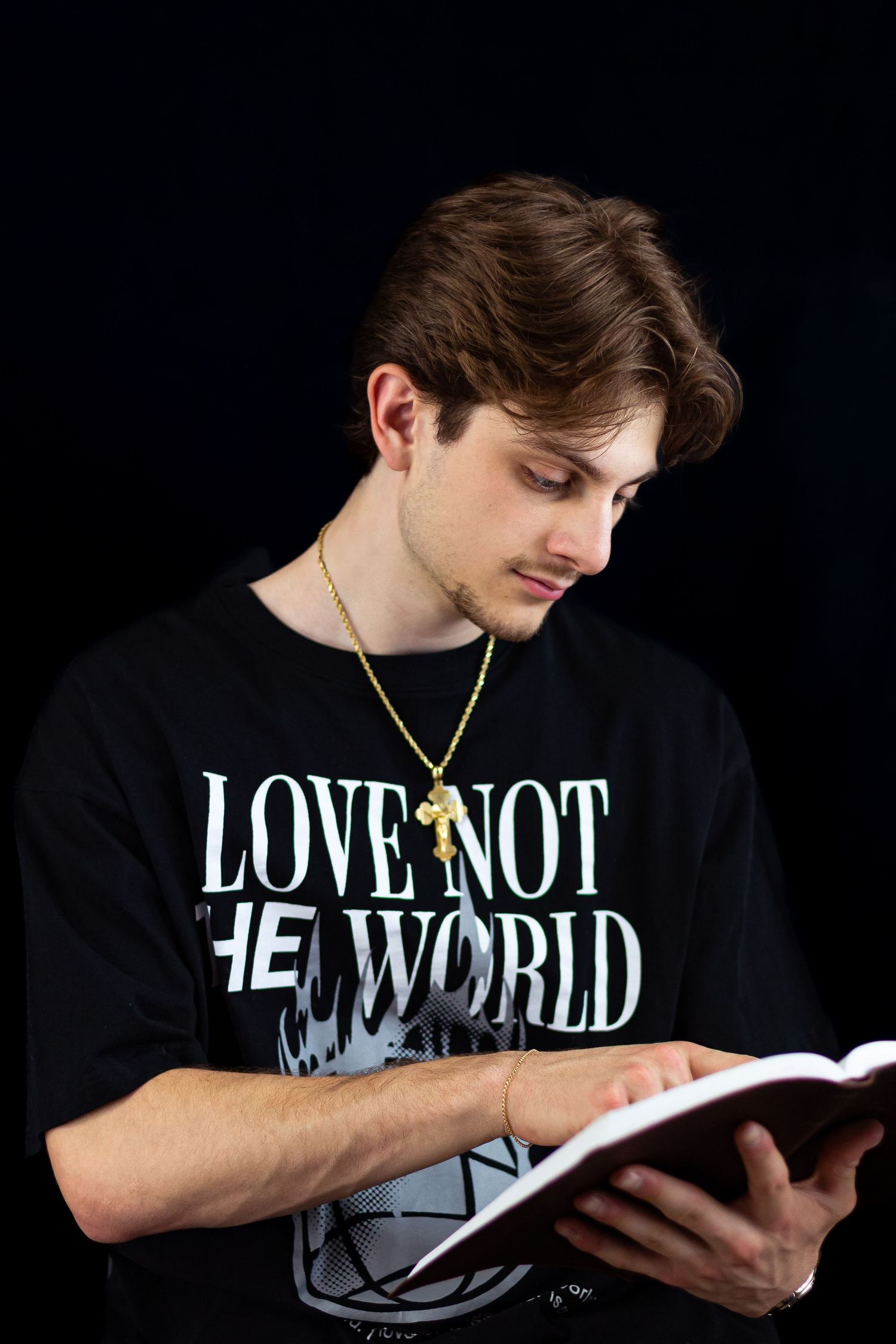 Young man with a gold cross necklace reads a Bible while wearing a black Love Not The World tee.