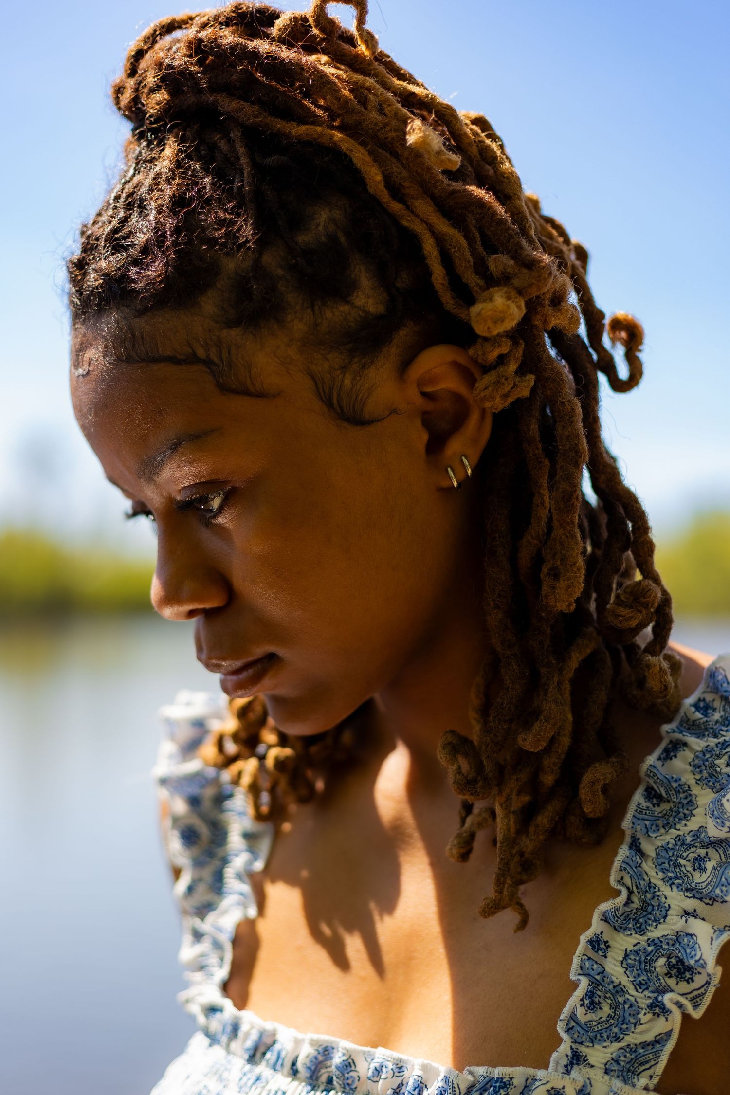 Young Black woman with brown locs gazing down near lakeside on clear sunny day.