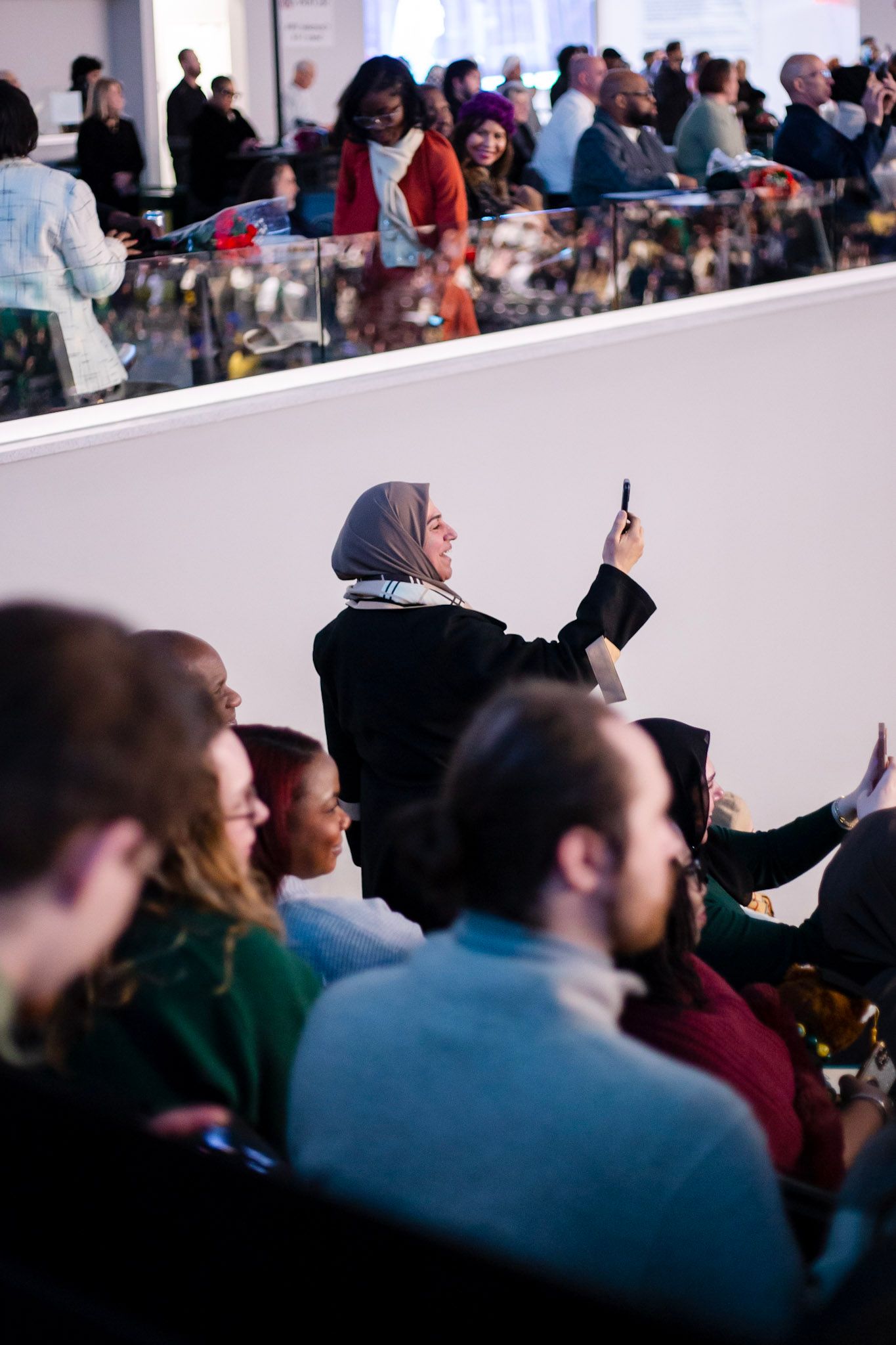 A woman in a hijab takes a selfie with her phone while seated in a crowded auditorium during an event.