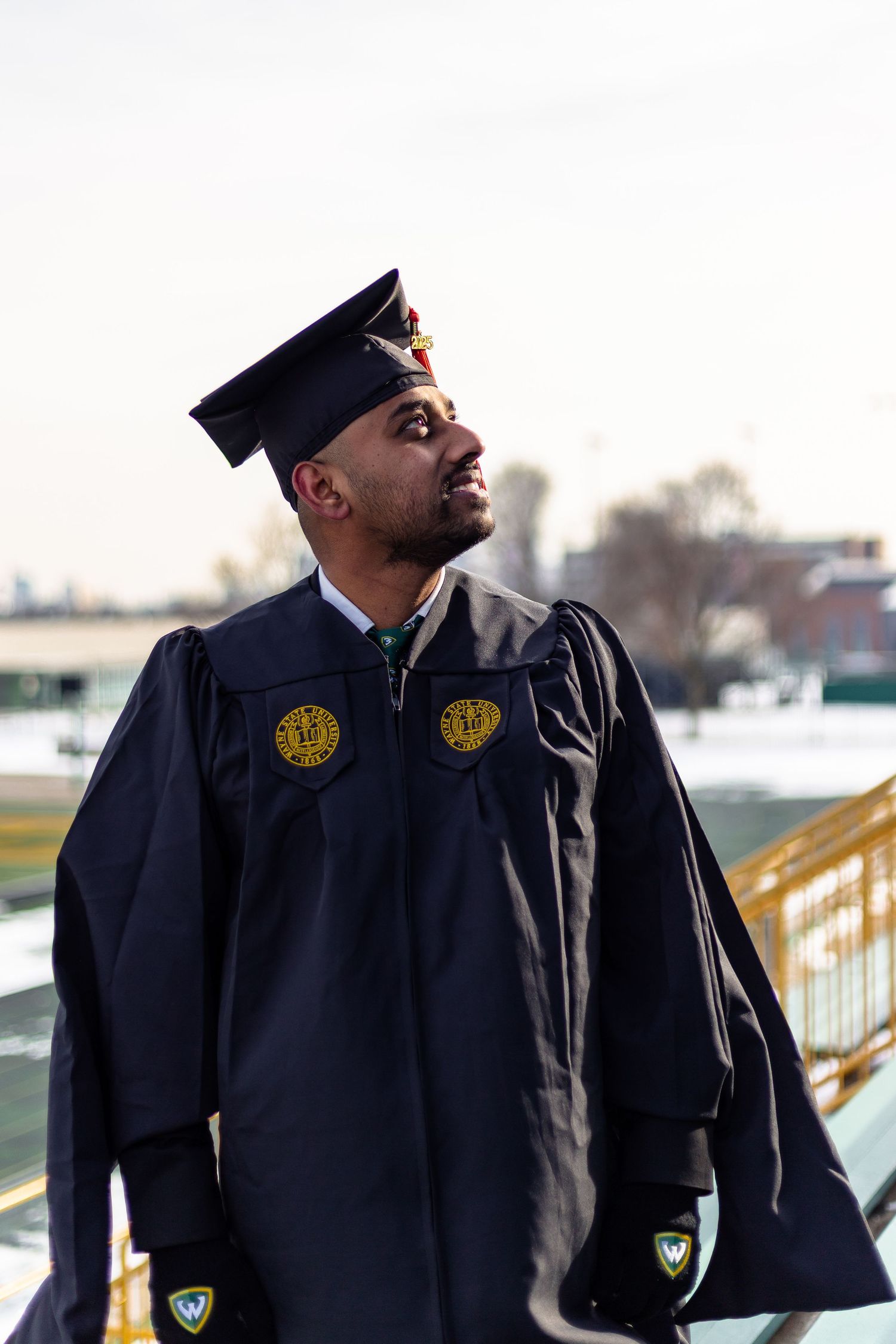 Wayne State University graduate in black regalia with gold patches looks up outdoors on campus.