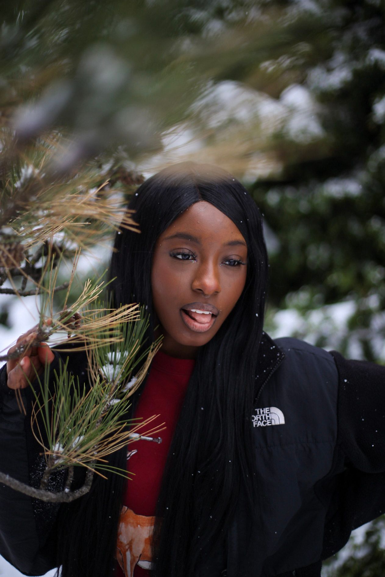 Young Black woman in North Face jacket holding snow-covered pine branch in winter forest setting.