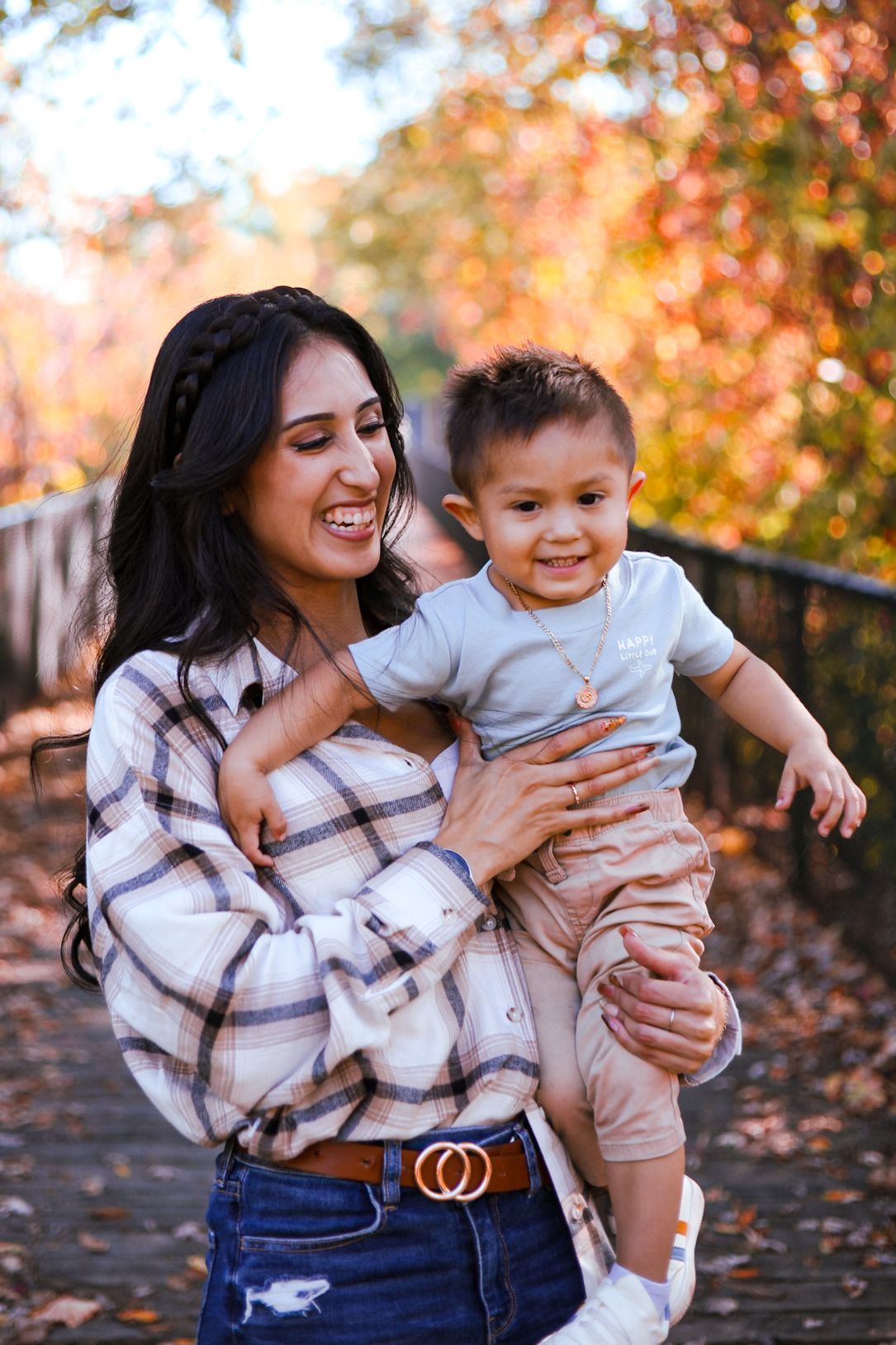 Joyful mother in plaid shirt holding her laughing toddler on a bridge in autumn park.